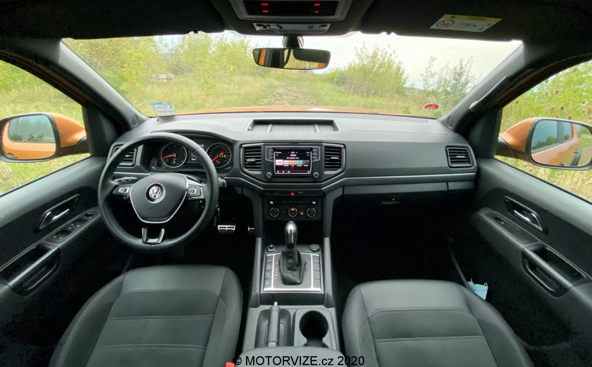 Imagem do interior da cabine dianteira do VW-Nutzfahrzeuge Amarok 2016. Tirada da perspectiva do banco traseiro, esta imagem mostra o volante central com controles montados, painel de instrumentos analógico com um pequeno display digital, sistema de infotainment no centro do painel com controles de áudio e clima, alavanca de câmbio indicando transmissão automática e bancos de couro. A imagem também captura o espelho retrovisor, espelhos retrovisores externos e vidros laterais dianteiros, destacando tanto as características tecnológicas como de conforto.