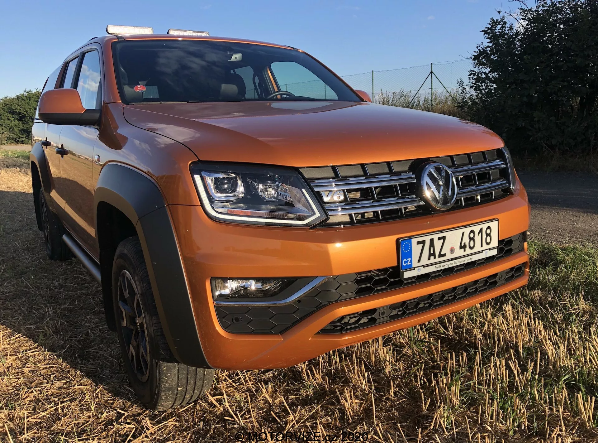 Imagem de um Volkswagen Amarok 2016 estacionado em uma área gramada com um céu claro ao fundo. Tirada de um ângulo frontal baixo inclinado para o lado do motorista, destaca a grade frontal com o logotipo da Volkswagen, faróis angulares, unidades de faróis de neblina na seção inferior preta do para-choque e o capô central elevado. O SUV, pintado em laranja metálico, possui guarnições de plástico preto nas rodas e uma placa de proteção no para-choque dianteiro. Também são visíveis os trilhos de teto prateados e as rodas de liga leve de cinco raios, enfatizando seu design robusto e pronto para off-road.