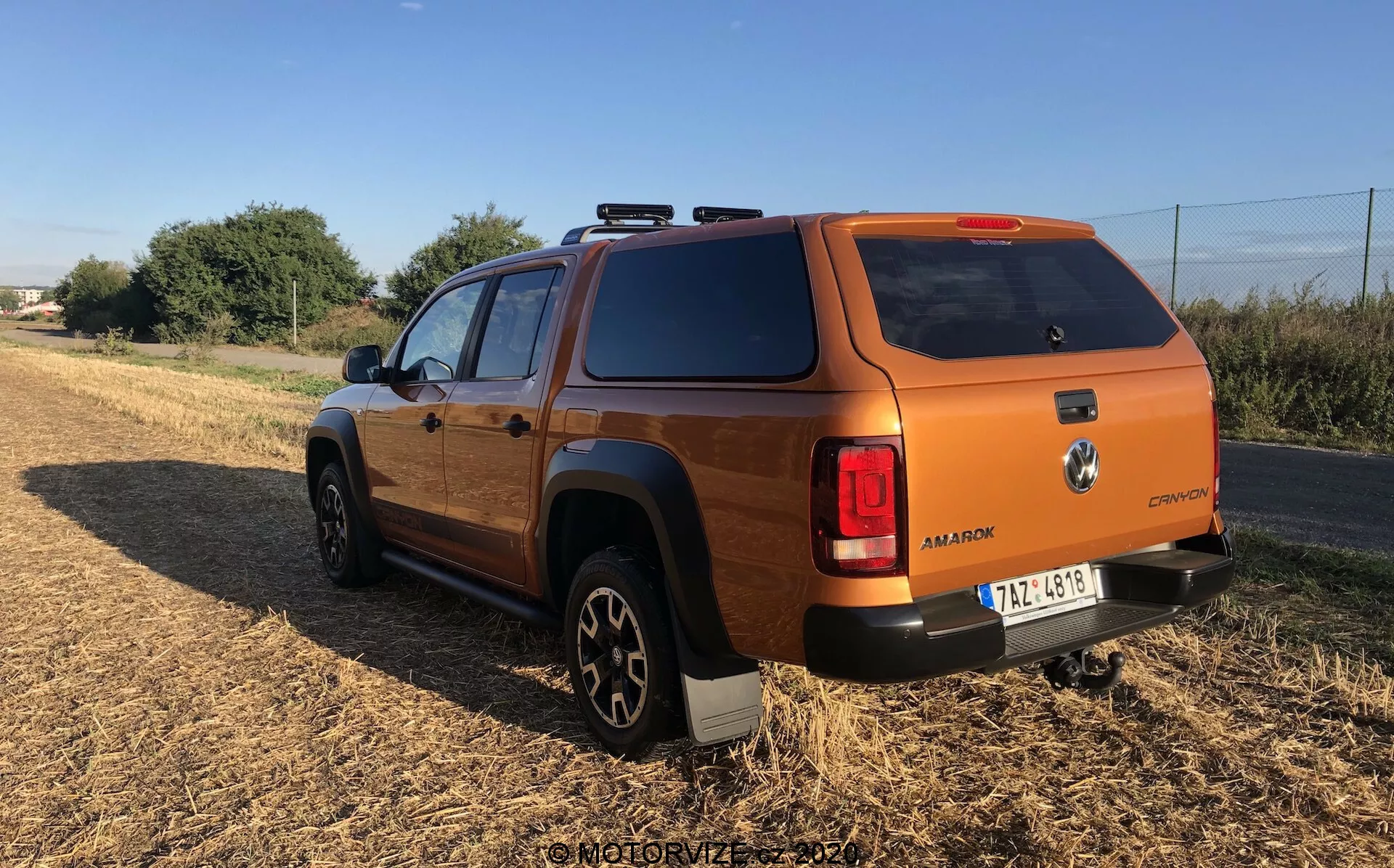 Vista traseira de um veículo utilitário moderno ou off-road de cor bronze ou laranja queimado, possivelmente um SUV médio ou picape, com uma tampa de compartimento de carga. O ângulo destaca a parte traseira e o perfil lateral, incluindo a roda traseira e a porta do passageiro, posicionado em piso não pavimentado com vegetação ao fundo. O veículo exibe um design robusto e quadrado típico de veículos off-road, com luzes traseiras envolventes, grandes pneus todo-terreno em rodas de liga preta, emblemas visíveis, trilhos no teto, vidros escurecidos na parte traseira e uma tampa sólida no compartimento de carga.