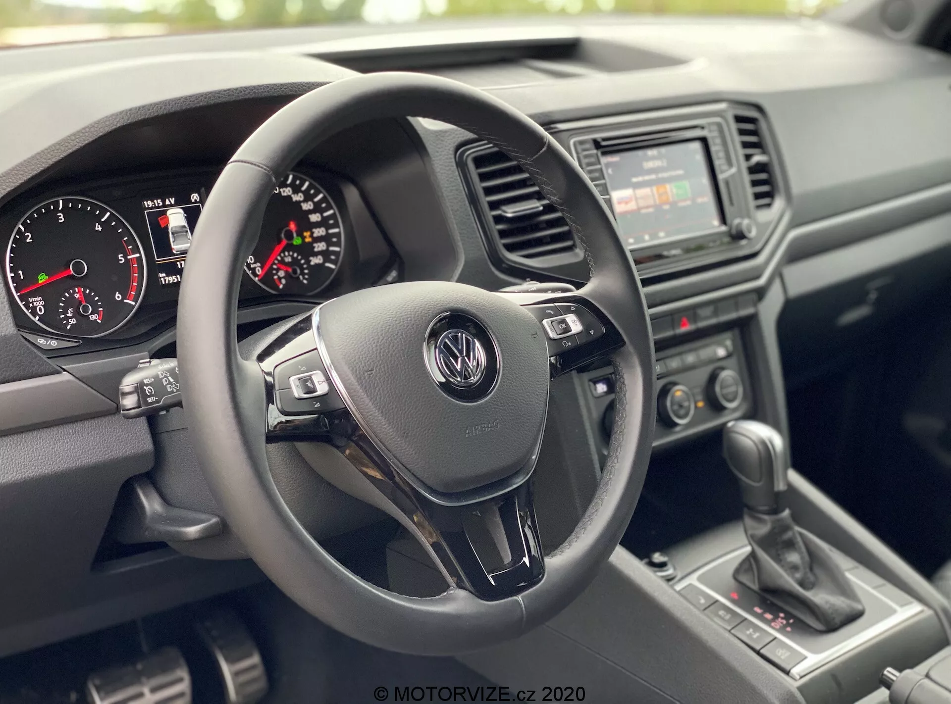 Vista interior do VW Nutzfahrzeuge Amarok 2016 da perspectiva do passageiro dianteiro, mostrando a área do cockpit do motorista. Foco central em um volante de três raios com logotipo da Volkswagen e botões de controle multifuncionais, painel de instrumentos com velocímetro analógico, tacômetro e um pequeno display digital, rádio básico com interruptores de controle climático manual no console central, alavanca de câmbio manual e materiais plásticos texturizados para o painel e portas, destacando um design durável e prático.
