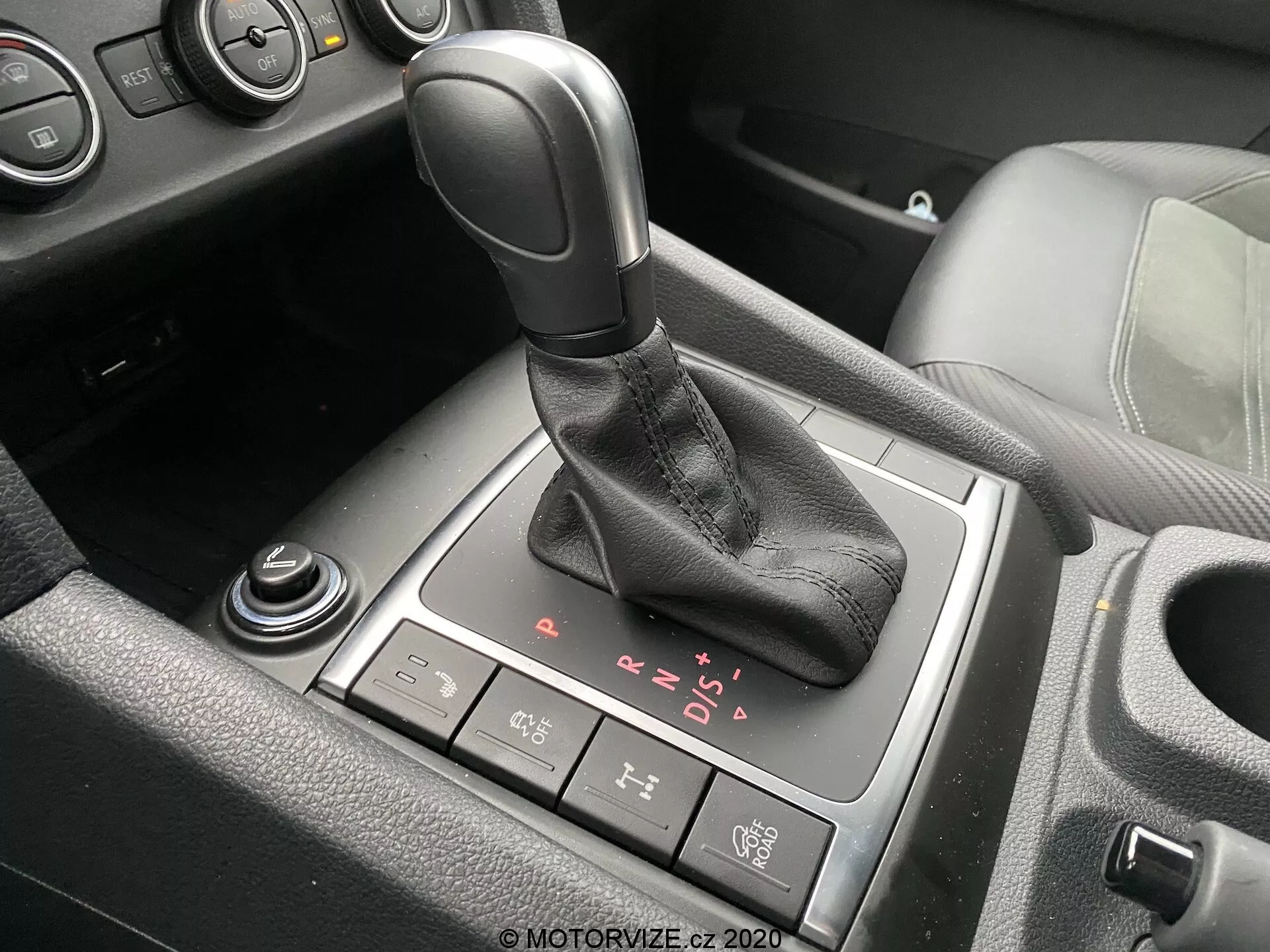 Vista detalhada da área da alavanca de câmbio no interior do VW Nutzfahrzeuge Amarok 2016. A alavanca de câmbio automática, com empunhadura na cor preta e coifa de couro, está posicionada centralmente na imagem com opções visíveis de P, R, N, D e modo sequencial manual. Um botão do freio de estacionamento eletrônico está localizado atrás da alavanca de câmbio, enquanto um dial rotativo parcialmente visível e outro dial estão situados à frente e na parte inferior esquerda, respectivamente. Vistas parciais do porta-copos e do console central com destaque em plástico preto e possivelmente em alumínio ou cromo também são visíveis, indicando um veículo moderno com acabamento interior de nível médio ou superior.