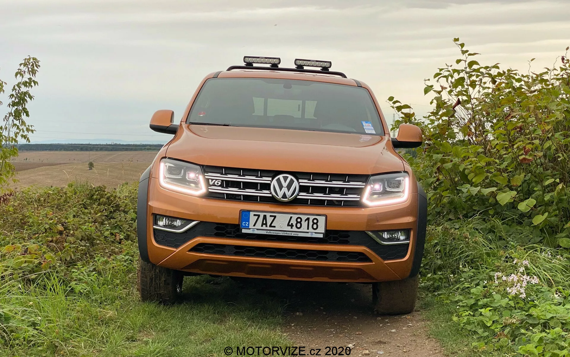 Vista frontal de um SUV Volkswagen Amarok laranja metálico estacionado na grama com fundo rural off-road, mostrando grade simétrica, faróis, acentos cromados e luzes auxiliares no teto.