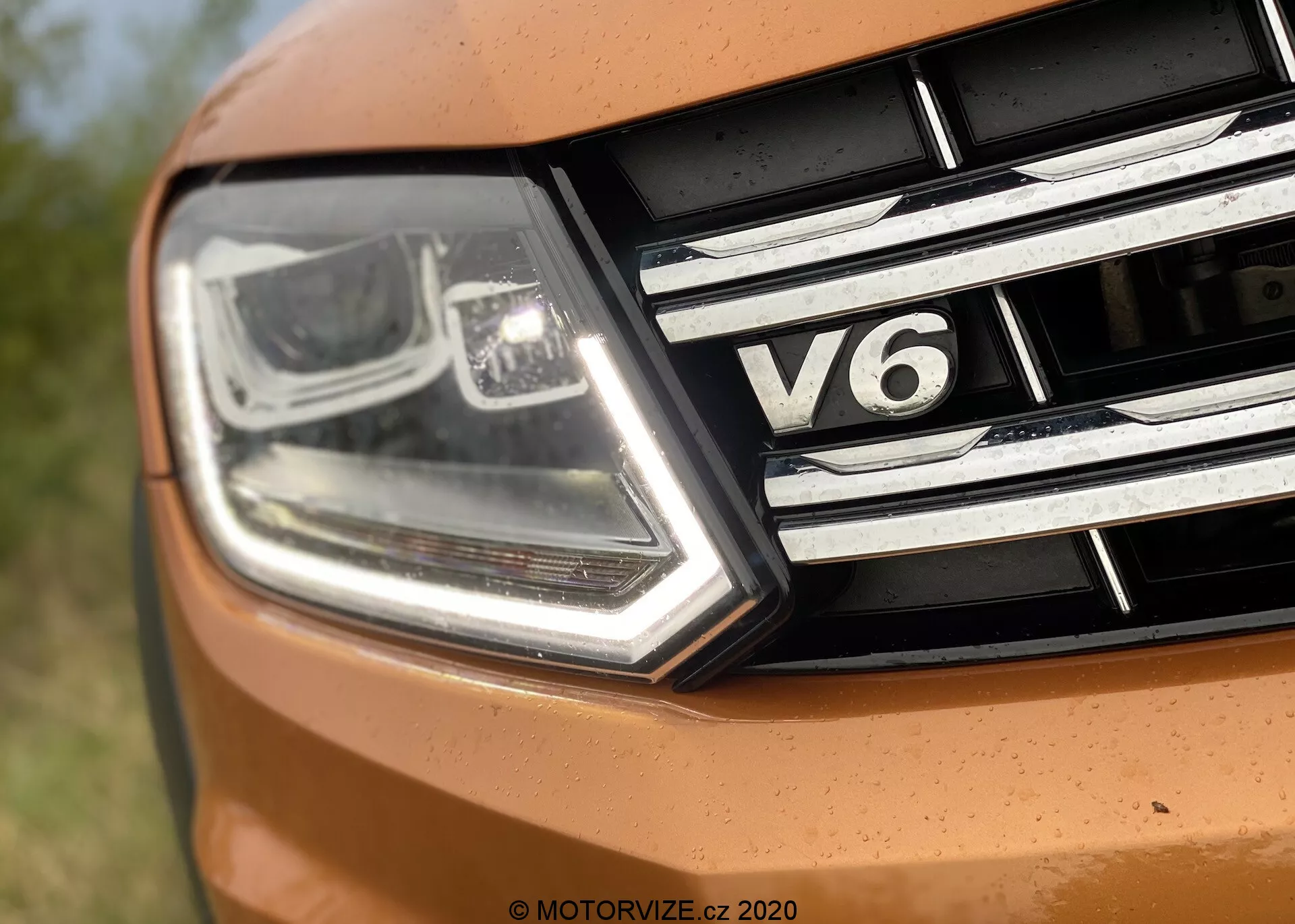 Close-up da seção frontal esquerda do VW Nutzfahrzeuge Amarok 2016. A imagem captura um farol moderno e angular com luzes diurnas de LED e tecnologia de feixe projetor, juntamente com detalhes cromados. A grade frontal possui tiras horizontais cromadas e um emblema 'V6', indicando o tipo de motor de seis cilindros do carro. Parte do capô aparece em um tom vibrante de laranja, sugerindo um design esportivo.