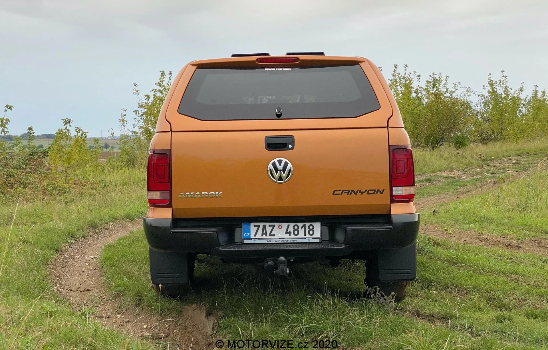 Vista traseira de uma picape Volkswagen Amarok 2016 laranja metálica estacionada ao ar livre em um caminho de terra irregular com grama em ambos os lados, sob um céu nublado. A foto mostra uma perspectiva clara da tampa traseira e dos recursos de design traseiro, incluindo o emblema Volkswagen, a placa com o nome Amarok e o emblema V6. O veículo possui um para-choque traseiro com degrau integrado, placa de registro centralizada e engate de reboque visível. As luzes traseiras verticais e a terceira luz de freio na linha do teto acima do para-brisa traseiro, bem como as linhas do descongelador, são visíveis.