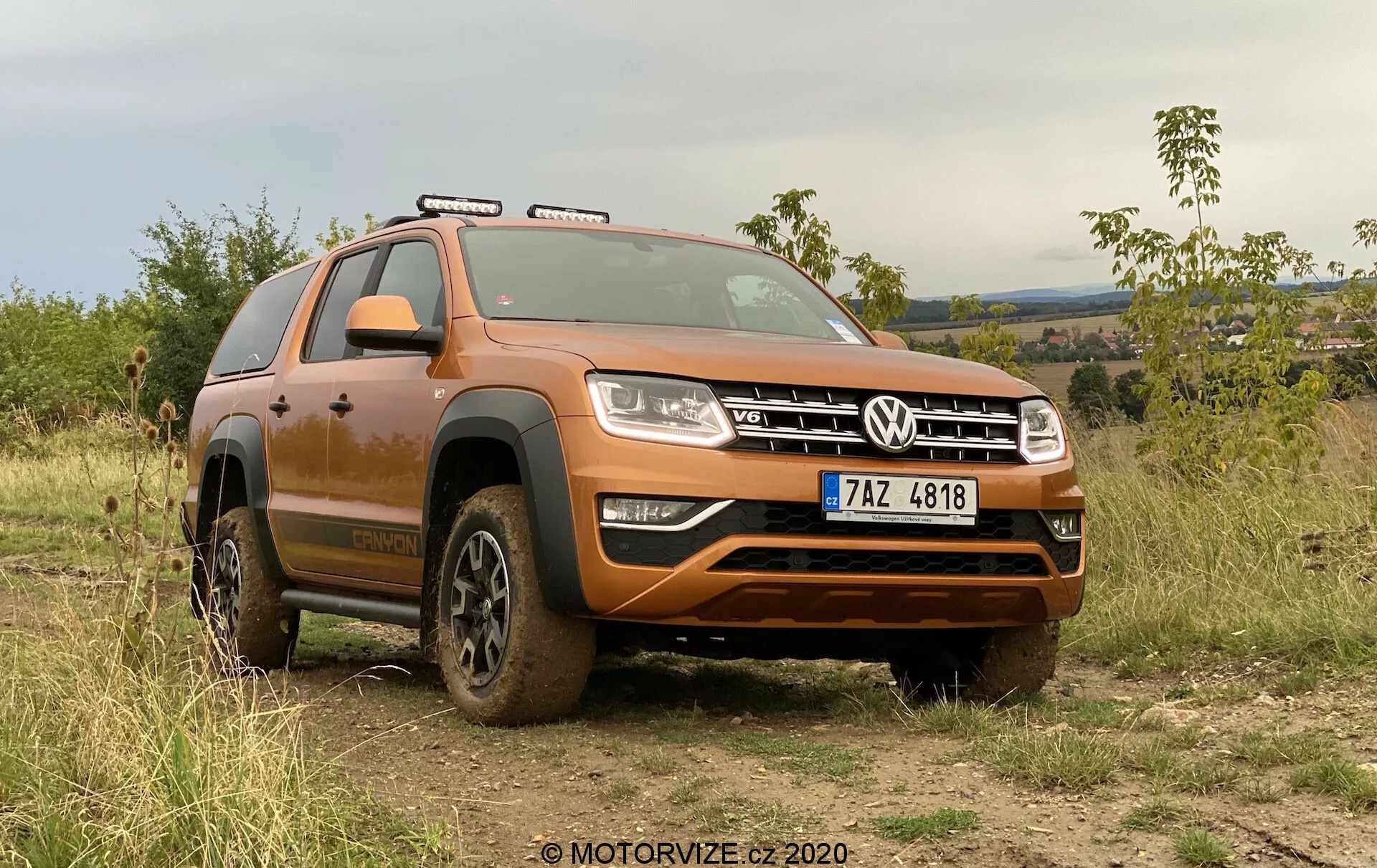 A imagem mostra um SUV moderno, modelo Volkswagen Amarok 2016, estacionado ao ar livre em terreno irregular, demonstrando sua capacidade de off-road. Visualizado de um ângulo frontal baixo, o quarto frontal do veículo é visível, coberto por resíduos lamacentos de uso off-road recente. A grade frontal larga com o emblema VW distintivo é ladeada por faróis retangulares com luzes diurnas LED. Abaixo da grade, um para-choque frontal robusto com uma placa protetora prateada está projetado para proteger o chassi. O capô contornado acrescenta uma aparência muscular ao SUV. Trilhos no teto são visíveis, indicando capacidade de carga adicional. As rodas, parcialmente cobertas de lama, têm pneus off-road com sulcos profundos para melhor tração em terrenos acidentados. Um arco proeminente sobre a roda dianteira e uma escada lateral são visíveis, facilitando a entrada e saída do veículo. Também são discerníveis as luzes no teto e os retrovisores laterais. O carro está posicionado contra um fundo de vegetação e terreno ondulado sob céu nublado, enfatizando sua capacidade off-road e apelo ao ar livre. O volante à esquerda e a placa visível sugerem um país com direção à direita. Esta imagem destaca o design do carro, características off-road e tecnologias avançadas de iluminação.