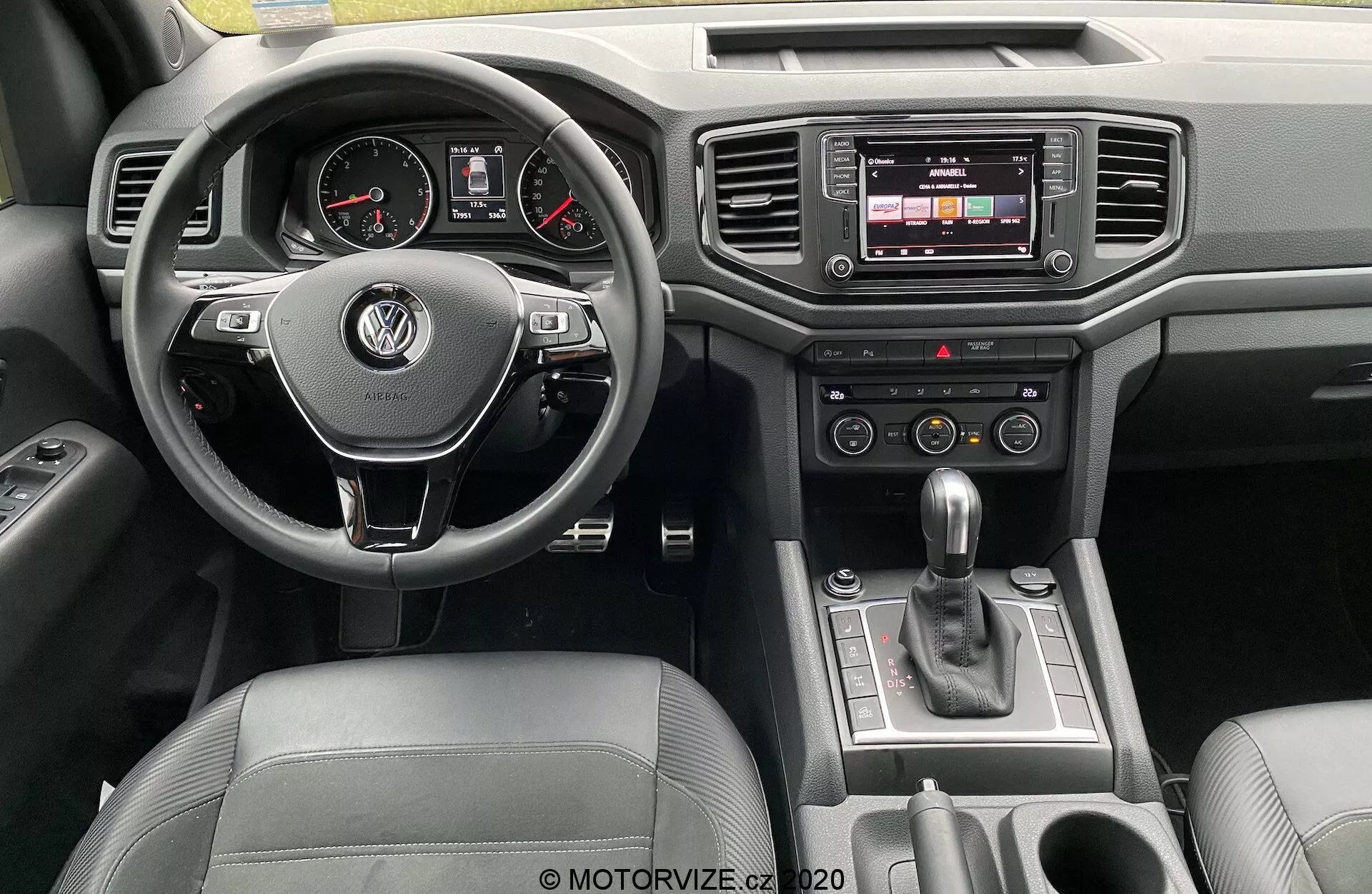 Vista do painel interior de um Volkswagen Amarok 2016 a partir da perspectiva do motorista. O volante multifuncional revestido em couro com o logotipo VW está centralizado, com mostradores analógicos e um display digital no painel de instrumentos. O console central apresenta um sistema de infoentretenimento com tela sensível ao toque, botões de controle de clima e alavanca de câmbio automática. Detalhes de luxo incluem acabamentos em couro e cromo, com controles de porta visíveis.