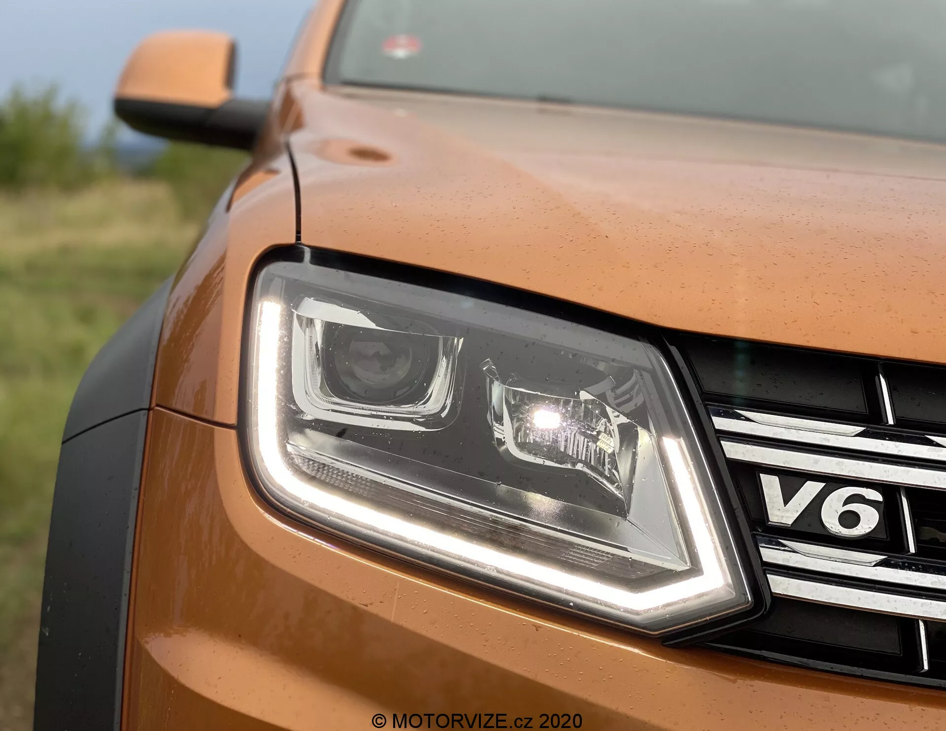 Close-up da visão parcial frontal do lado esquerdo dianteiro de um VW Nutzfahrzeuge Amarok 2016 de cor bronze ou cobre. A imagem mostra o design dos faróis LED com luzes diurnas distintas, uma grade cromada com emblema 'V6', painel da carroçaria com pintura metálica, borda contornada do capô e marca d'água 'MOTORVIZE.CZ 2020', sugerindo origem de um site de avaliações tcheco.