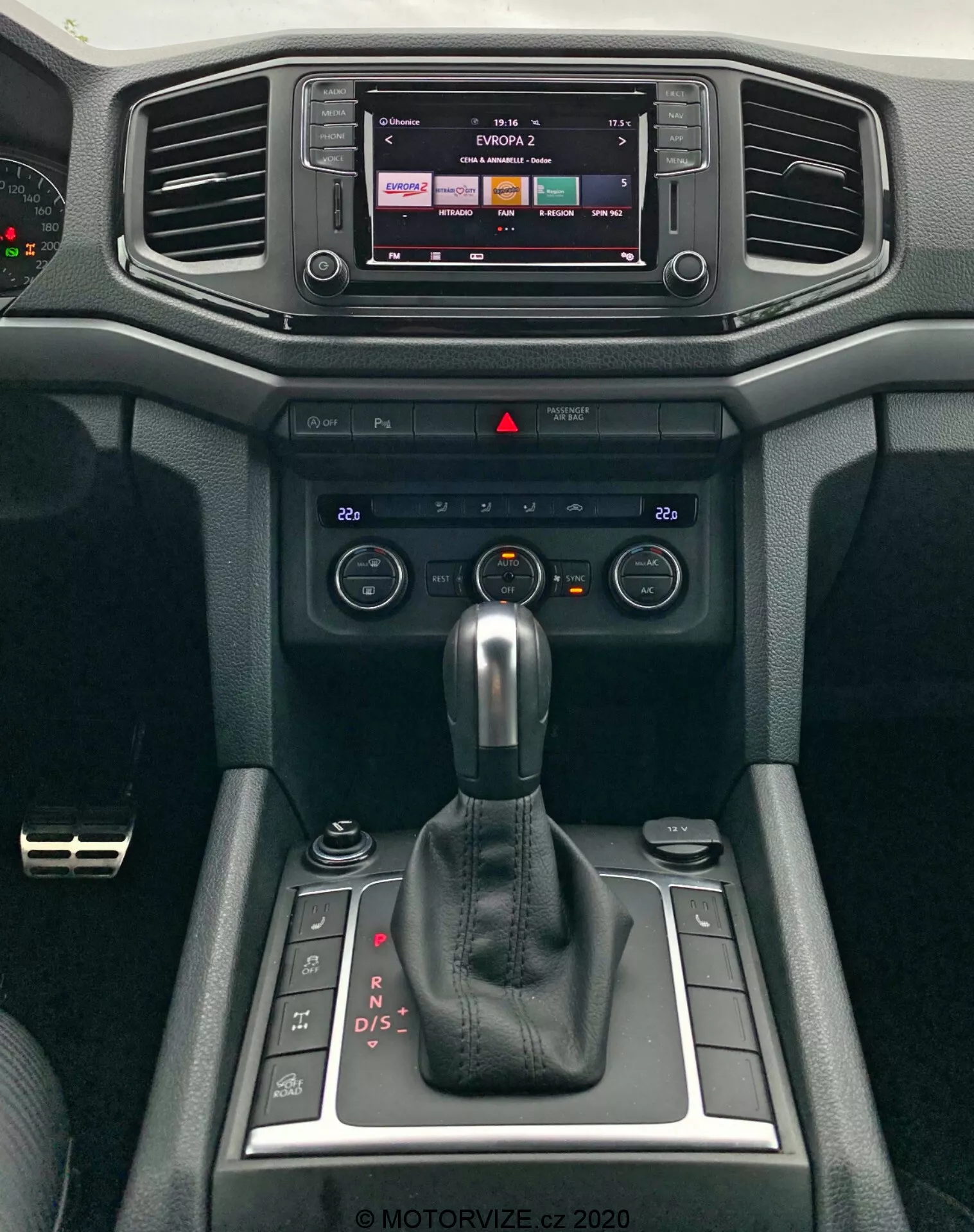 Close-up da moderna consola central do VW Amarok 2016, destacando o sistema de infotainment e alavanca de câmbio. O display do infotainment sugere capacidades de tela sensível ao toque, provavelmente mostrando saída de rádio ou mídia. Abaixo, estão saídas de ar horizontais cercadas por materiais suaves ao toque com design texturizado. Os controles de clima e interruptores estão posicionados abaixo das saídas de ar para ajuste de temperatura e fluxo de ar. Uma alavanca automática de câmbio em modo 'P' domina o centro, indicando a transmissão automática do veículo com opções de mudança manual. Pequenos compartimentos estão ao lado da alavanca de câmbio, junto com um botão rotativo, provavelmente para controle de infotainment ou modos de direção. O design interior enfatiza tanto a funcionalidade quanto o conforto.