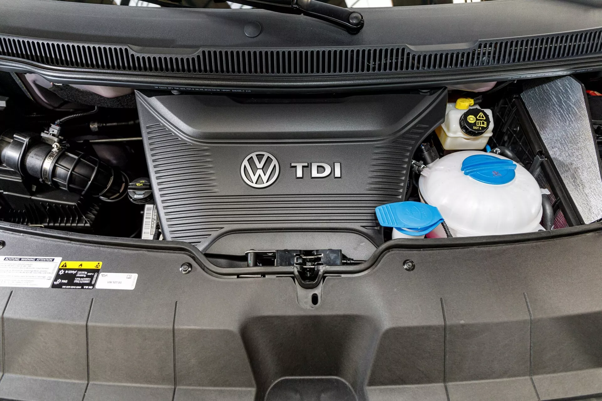 Vista detalhada do compartimento do motor a diesel do VW Nutzfahrzeuge Transporter 2019 (T6.1 California) mostrando a tampa do motor TDI com o logo da Volkswagen, reservatório de refrigerante e bateria do carro