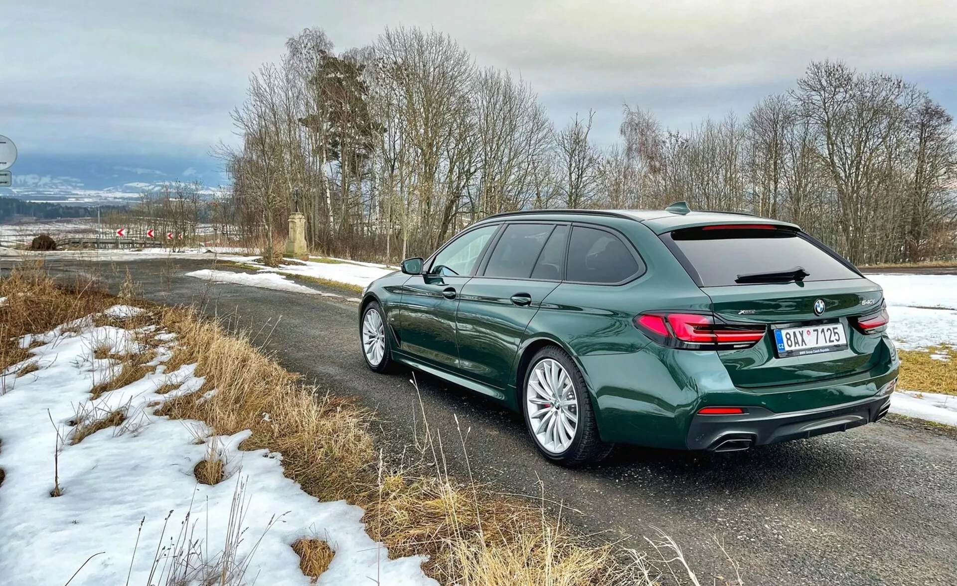 Vista traseira três quartos de uma carrinha BMW, mostrando a parte traseira do veículo com luzes traseiras em forma de L, perfil lateral esquerdo incluindo portas e a curva do teto com barras de teto, jantes de liga leve com pneus para estrada, em pintura verde escuro metálica, estacionada numa estrada de campo com leve neve, destacando elementos de luxo e design moderno.