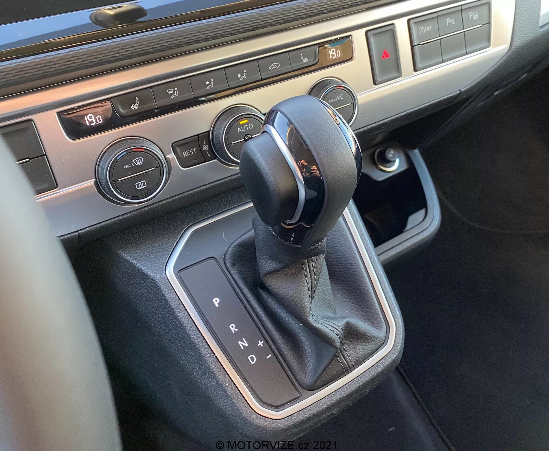 Vista aproximada do console central do VW Nutzfahrzeuge T6-1 Multivan 2019, focada na área da alavanca de câmbio. A imagem mostra um seletor de transmissão automática com costura de couro, cercado por acabamento metálico, botões de controle de clima com display de temperatura e controles de aquecimento de assento, destacando a tecnologia interior moderna e os materiais premium do veículo.