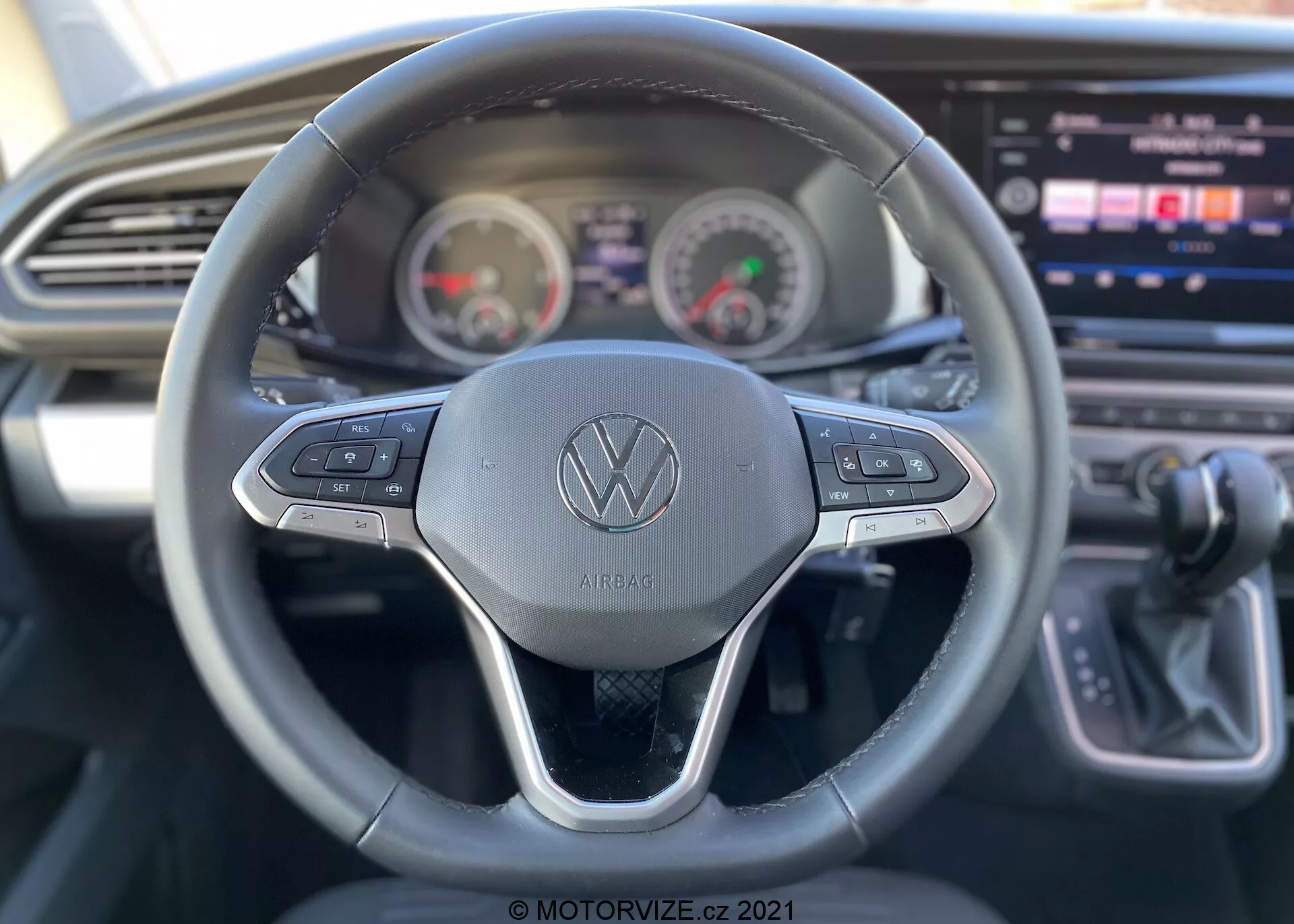 Vista interior de um VW Nutzfahrzeuge Transporter 2019 (T6-1 Multivan) focada na perspectiva do condutor. Vista detalhada do volante com o logótipo Volkswagen e botões de controlo para várias funções como o controlo de cruzeiro e a navegação multimédia. Atrás do volante, o painel de instrumentos mostra um velocímetro e um tacómetro em estilo analógico com um ecrã digital no centro. Consola central parcialmente visível com um grande ecrã tátil de informação e entretenimento, juntamente com um design elegante do painel de instrumentos com ventilação horizontal. Manípulo de mudanças à direita indicando transmissão automática. A imagem destaca funcionalidades de usabilidade com reduzida profundidade de campo focada principalmente no volante.