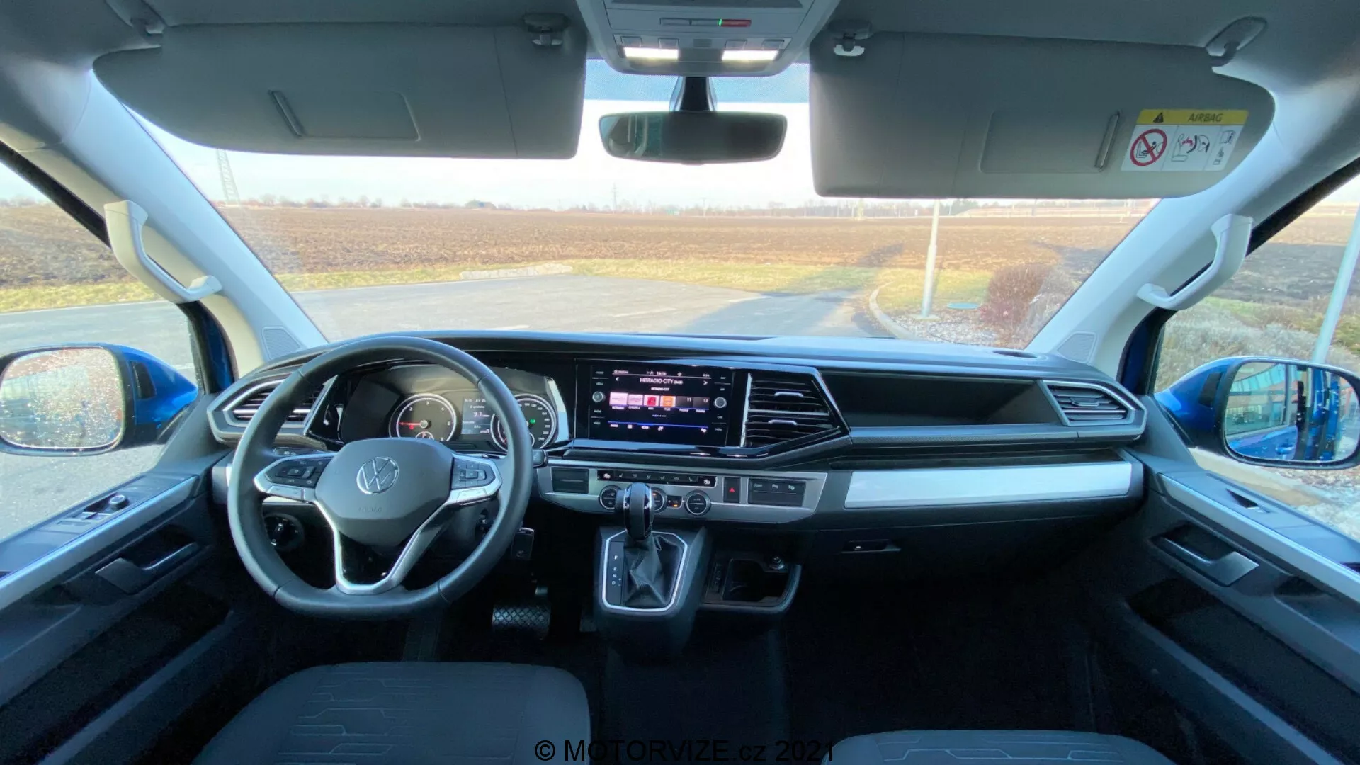 Interior de um VW Nutzfahrzeuge Transporter 2019 (T6.1 Multivan), capturado a partir da área dos assentos traseiros em direção ao painel frontal. A foto foi tirada durante o dia com luz natural entrando pelo para-brisa dianteiro, proporcionando uma visão detalhada do espaço da cabine, dos assentos dianteiros, do volante com controles montados, da tela de toque central do sistema de infotainment, dos botões e manípulos do HVAC, da alavanca de câmbio automática, dos espaços de armazenamento, das portas de conectividade, dos controles de porta e espelho, dos recursos de segurança e do design confortável dos assentos.