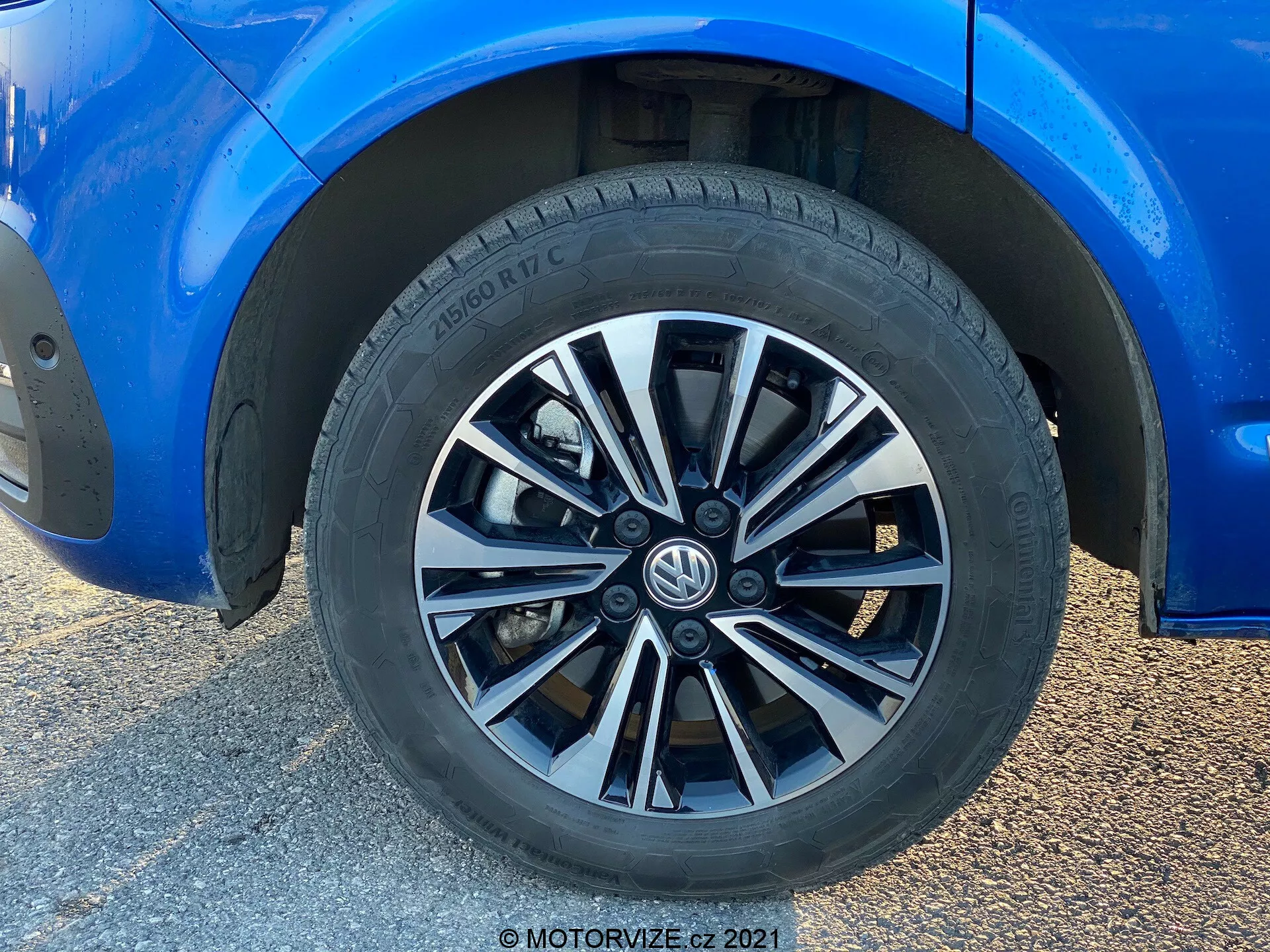 Vista em close-up de uma roda do Volkswagen T6-1 Multivan 2019. Este tiro em quadro inteiro apresenta um design de roda multi-raios com acabamentos alternados em prata e escuro. O pneu, da marca Hankook, revela um padrão de piso para desempenho em rua e especificações parciais do pneu. A carroceria azul circundante, em ótimo estado, exibe um brilho reflexivo. Indica uma distância ao solo moderada e pinças de freio padrão parcialmente visíveis. A luz natural realça a composição focada na roda, com o fundo desfocado devido a uma tomada em ângulo baixo.