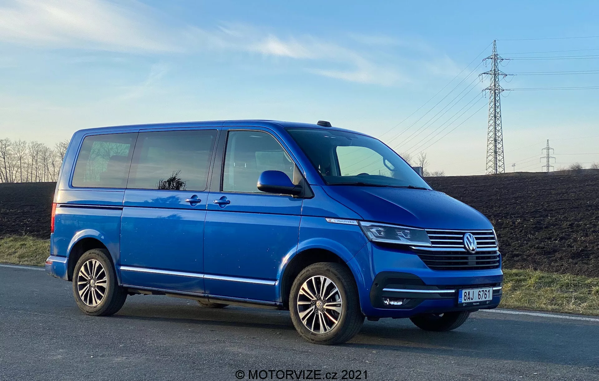 Uma van Volkswagen Transporter T6 azul, mostrando seu perfil lateral de um ângulo baixo, enfatizando seu comprimento e altura. A imagem apresenta detalhes como a porta deslizante do passageiro, a porta dianteira do passageiro, a roda traseira do lado do passageiro, aros de liga leve com vários raios e vistas parciais do farol, grade Volkswagen com emblema e para-choque dianteiro. A foto, tirada no final da tarde, mostra a distância do solo e reflete o céu e os arredores nas janelas, com céu claro e linhas de energia ao fundo.