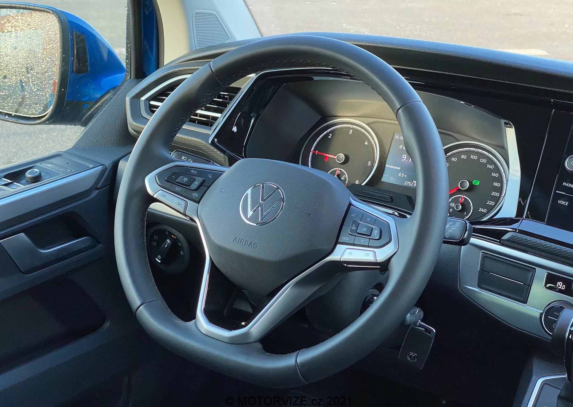 Vista interior do VW Nutzfahrzeuge Transporter 2019 (T6-1 Multivan) a partir da perspectiva do motorista, focando no volante e no painel de instrumentos. O volante apresenta o emblema Volkswagen e é equipado com revestimento de couro, botões multifuncionais e paddle shifters. O painel de instrumentos parcialmente visível inclui mostradores analógicos e uma tela digital. A tela central de infotainment e os controles de climatização estão ligeiramente visíveis, indicando características modernas de multimídia e conforto.