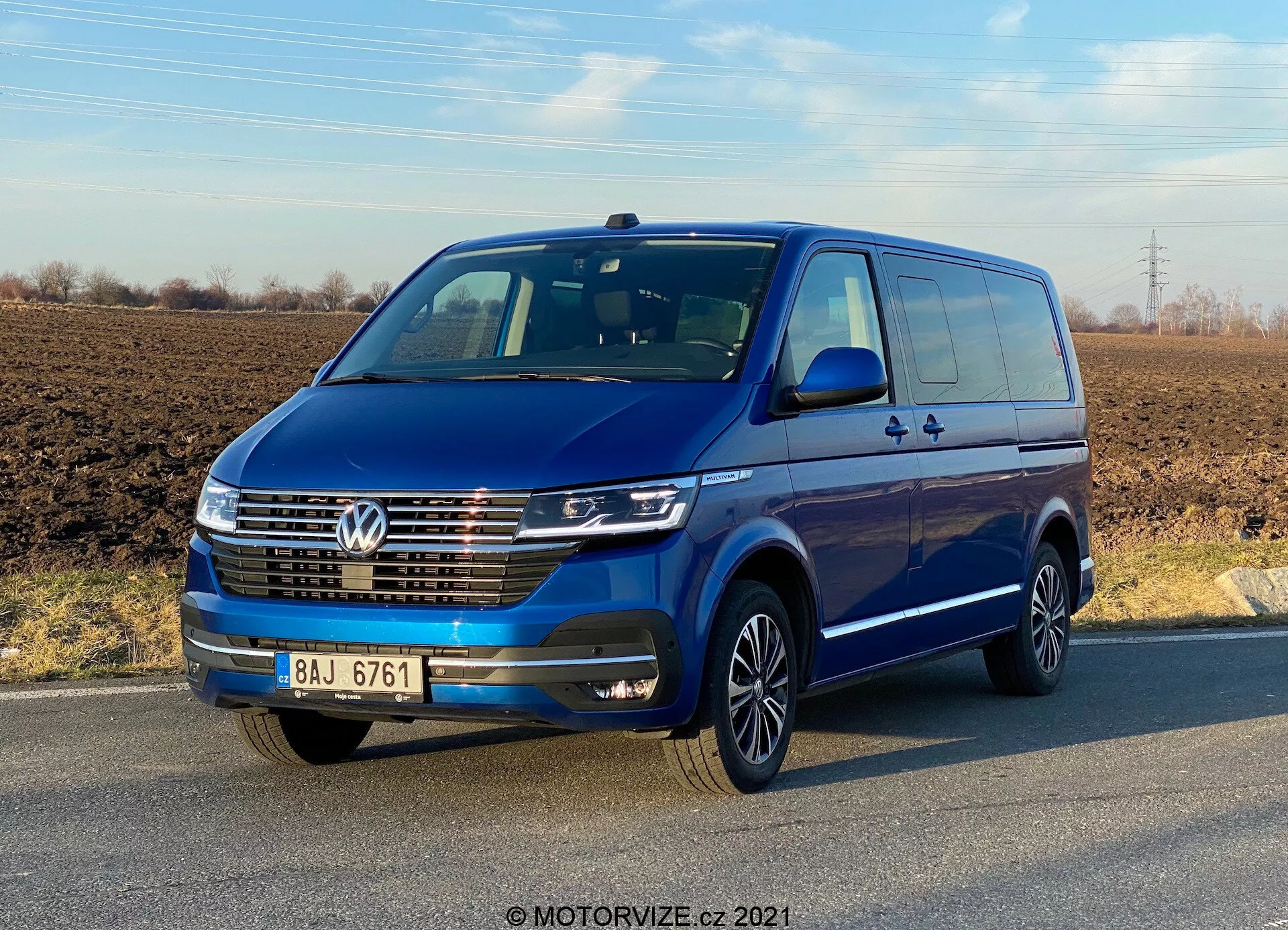Vista frontal de três quartos de um Volkswagen Transporter T6.1 Multivan azul de 2019 sob um céu claro. O veículo apresenta um emblema da Volkswagen na grade, luzes diurnas LED, acabamento cromado na grade frontal e rodas de liga leve de múltiplos raios. Espelhos e para-choques na cor do veículo sugerem um modelo de alta especificação. As portas e alças das portas, incluindo uma porta traseira deslizante, são visíveis, indicando sua capacidade para vários passageiros ou carga.