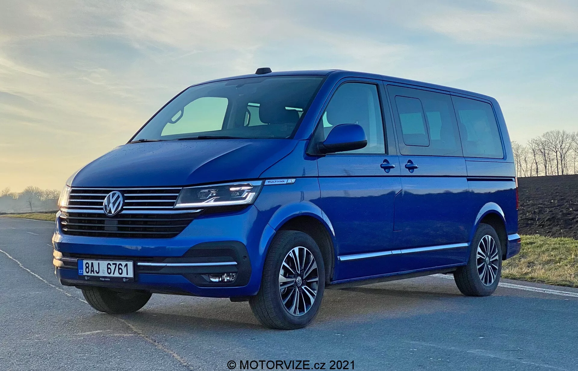 Imagem de uma van azul Volkswagen Transporter T6.1 Multivan de 2019, fotografada de um ângulo baixo para acentuar sua estatura e tamanho. A vista frontal de três quartos mostra o design frontal e lateral do veículo, incluindo a distinta grade Volkswagen com linhas horizontais, o emblema central, faróis modernos que provavelmente incluem tecnologia LED ou Xenon e um pára-choques dianteiro elegante com uma ampla entrada de ar. Os espelhos laterais aerodinâmicos com indicadores de direção integrados, amplo espaço das janelas com uma porta lateral deslizante, rodas de liga leve com pneus de perfil baixo e uma iluminação suave destacam a praticidade e o apelo estético da van. A marca d'água 'MOTORVIZE.cz 2021' sugere que a imagem é de 2021.