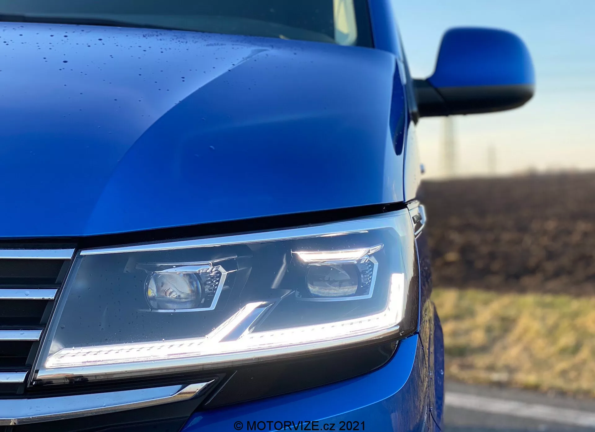 Uma foto em close-up e em ângulo baixo da seção frontal esquerda do VW Nutzfahrzeuge Transporter 2019 (T6.1 Multivan) azul, destacando os faróis modernos em LED de formato angulado com luzes diurnas, parte da grade com detalhes cromados e uma visão do contorno do capô e espelho lateral. A foto, tirada com baixa profundidade de campo, dá ênfase aos faróis e à carroceria circundante, destacando a tecnologia avançada de iluminação, o design contemporâneo e a qualidade da pintura, complementada por gotas de chuva indicando uma camada exterior detalhada.