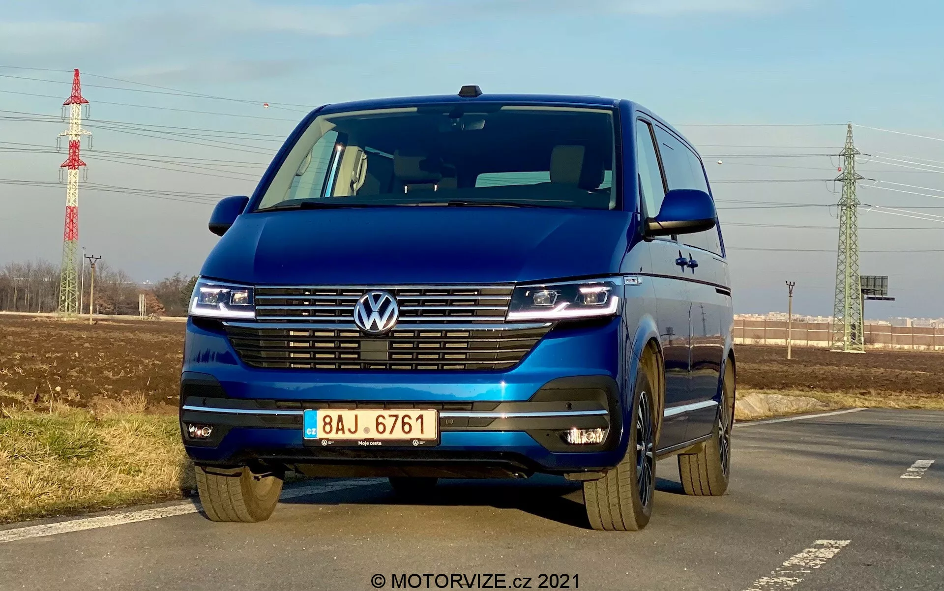 Volkswagen T6-1 Multivan 2019 azul capturado de um ângulo frontal baixo destacando a grade, faróis de LED com luzes diurnas, emblema Volkswagen, grade larga, para-choques bicolor e rodas de liga leve; estacionado ao lado de uma estrada com céu claro, folhagem distante, poste de energia e infraestrutura ao fundo.