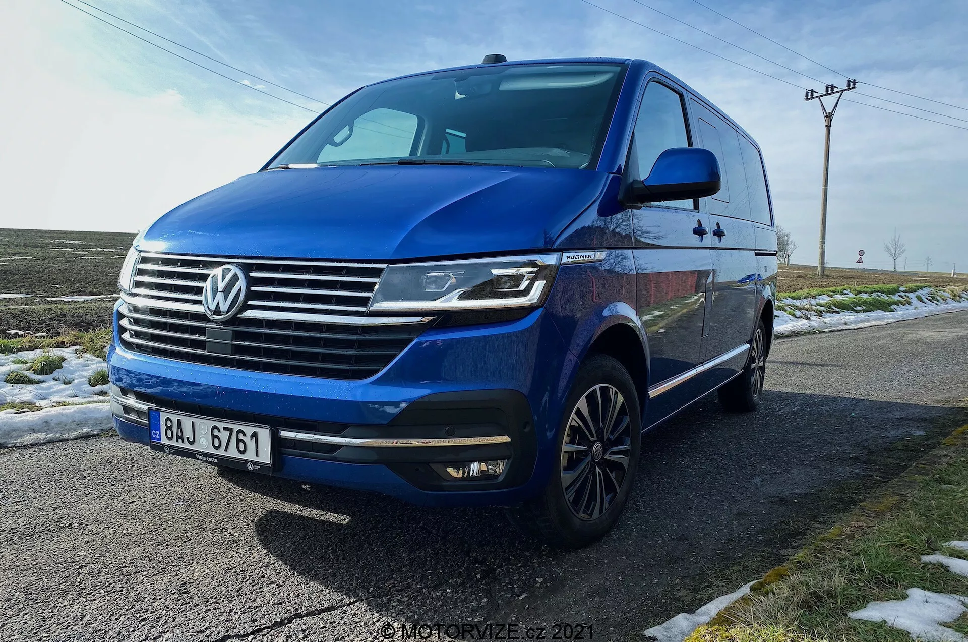 Volkswagen T6.1 Multivan Transporter 2019 azul capturado de um ângulo frontal a três quartos baixo, mostrando a parte frontal do carro, grelha com logótipo Volkswagen proeminente, barras horizontais cromadas, faróis LED angulares com luzes diurnas, para-choques dianteiro com luzes de nevoeiro integradas e entradas de ar, linha dinâmica que percorre os lados, rodas de liga leve multiraios, sinais de mudança de direção integrados nos espelhos laterais, janelas escurecidas. O veículo está estacionado numa estrada molhada ao lado de relva com neve, com um fundo rural que inclui postes de serviços públicos e céu aberto.