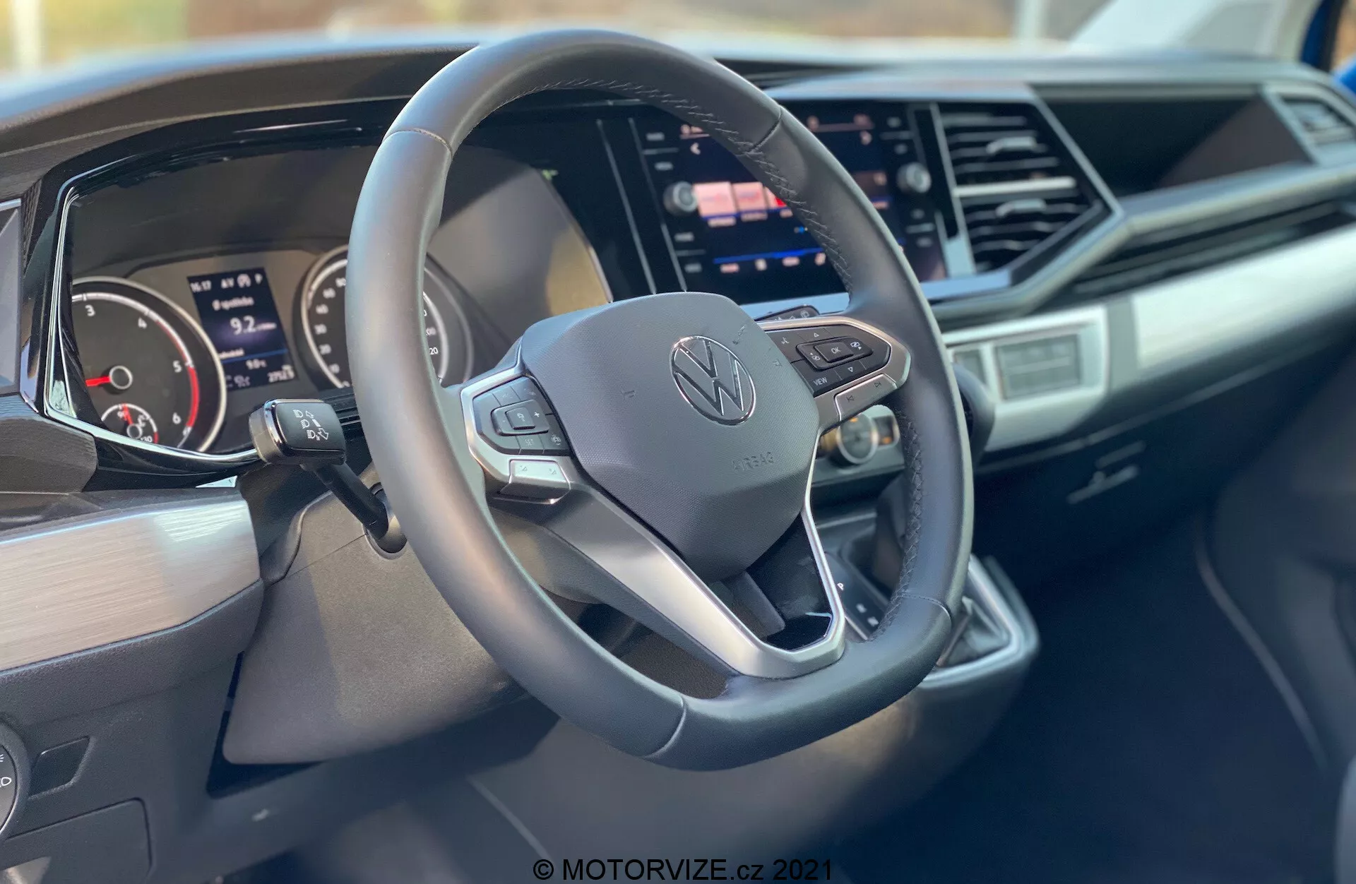 Imagem do interior de um Volkswagen Nutzfahrzeuge Transporter 2019 (T6-1 Multivan) focando no volante e no painel de instrumentos. O close-up, tirado do lado do passageiro, destaca um volante de três raios com fundo plano e botões de controle com o logotipo central da Volkswagen, um painel de instrumentos digital, uma grande tela tátil central de infoentretenimento, design elegante do painel com acentos angulares e marca no lado do passageiro do painel.