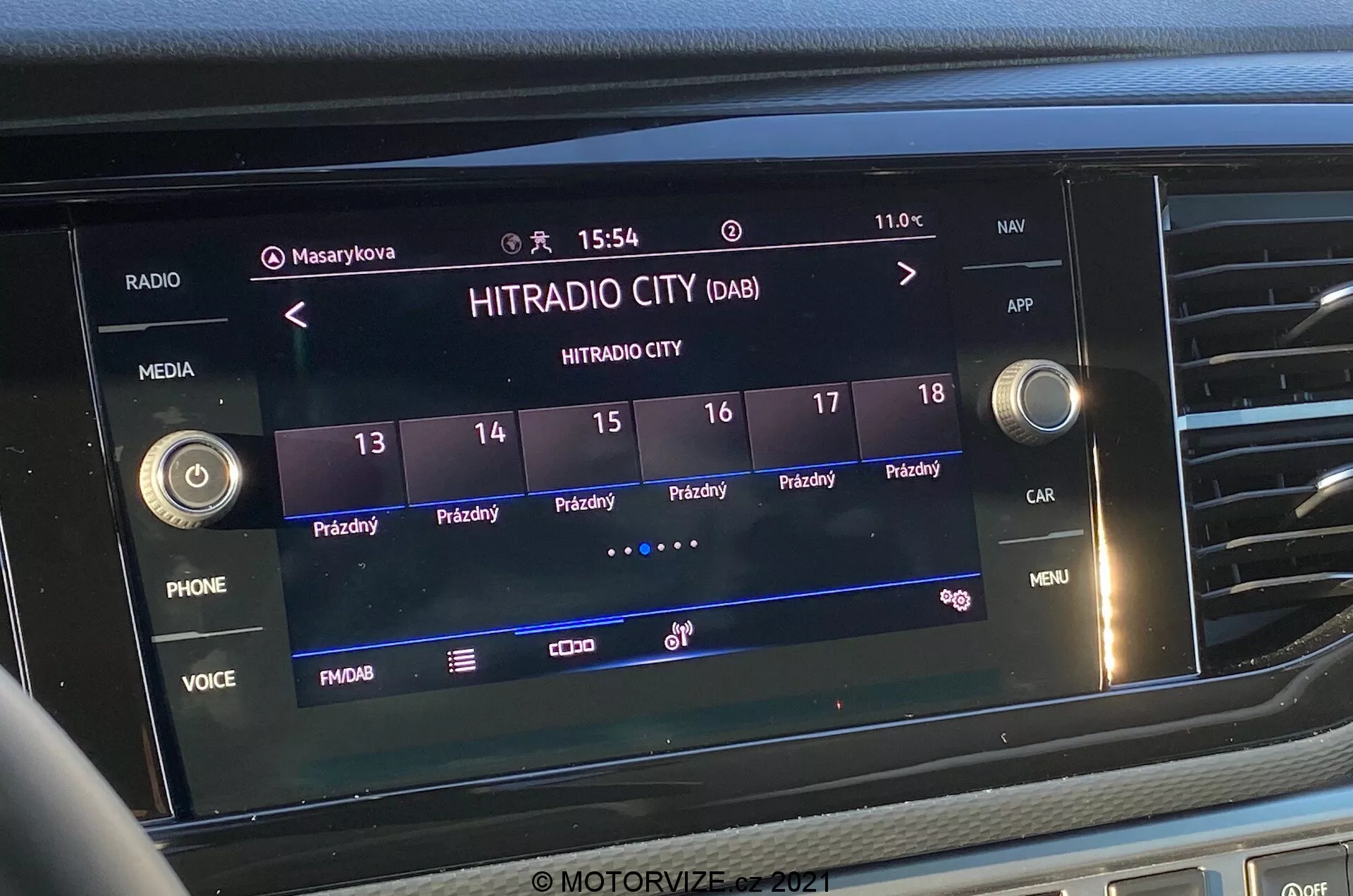 Foto frontal close-up da tela do sistema de infoentretenimento no VW-Nutzfahrzeuge Transporter 2019 (T6-1 Multivan). A imagem destaca a estação de rádio digital 'HITRADIO CITY' selecionada, com várias opções ativadas por toque e botões de função como 'PHONE', 'VOICE' e 'MENU'. A tela mostra a hora '15:54' e a temperatura externa '11.0°C', com ícones para perfis de usuário, configurações do veículo e intensidade do sinal. A tela está integrada na porção central do painel, cercada por botões táteis e botões de controle.