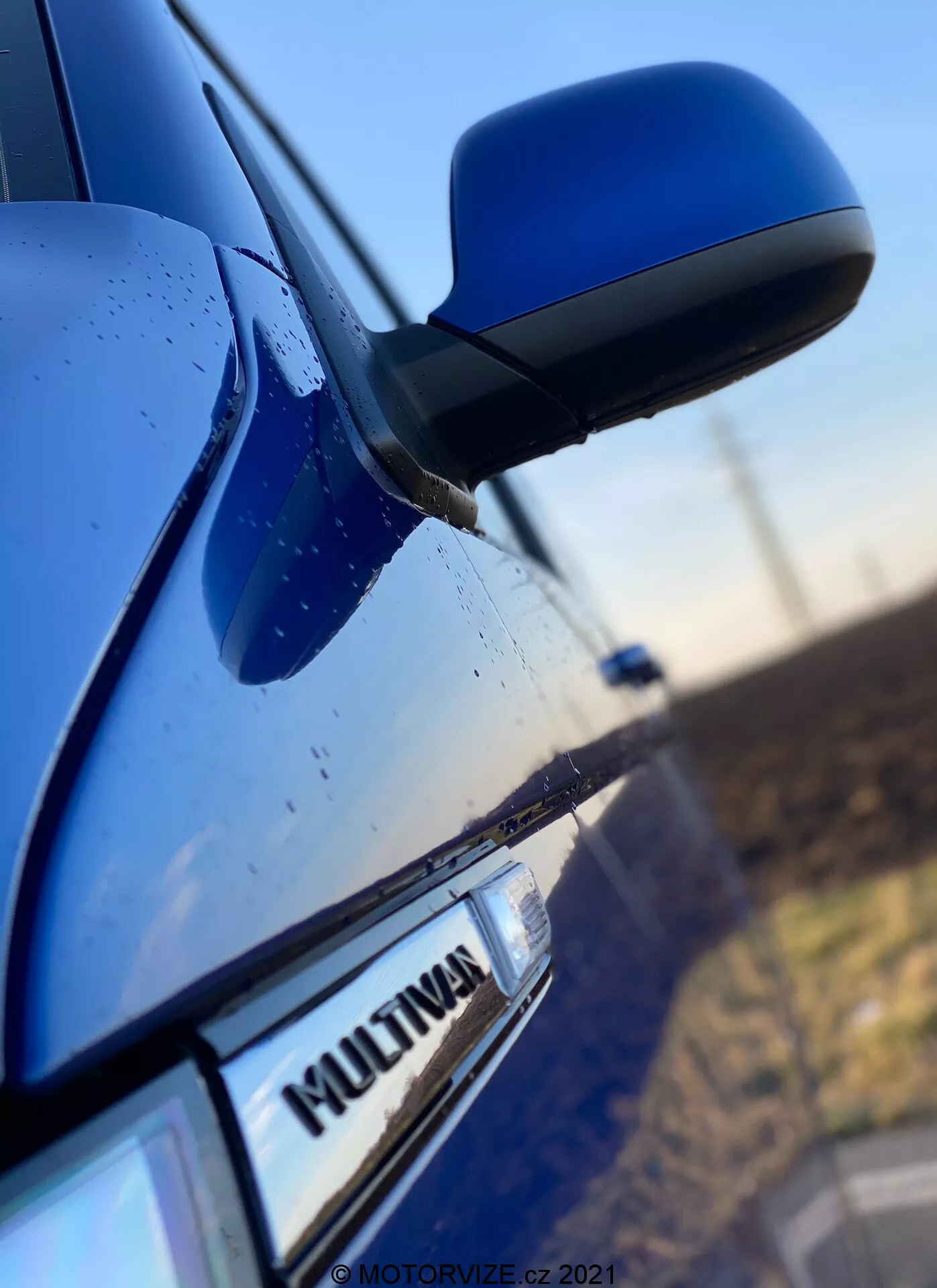 Foto em close-up da lateral de um VW Nutzfahrzeuge Transporter 2019 azul (T6-1-Multivan) mostrando parte da porta do passageiro dianteiro, o espelho lateral e uma parte da porta traseira. O foco está no emblema 'MULTIVAN' do carro, com o espelho lateral aerodinâmico ligeiramente desfocado. A carcaça do espelho combina com a cor da carroceria, e gotas de água visíveis na superfície sugerem características de impermeabilidade ou chuva recente.