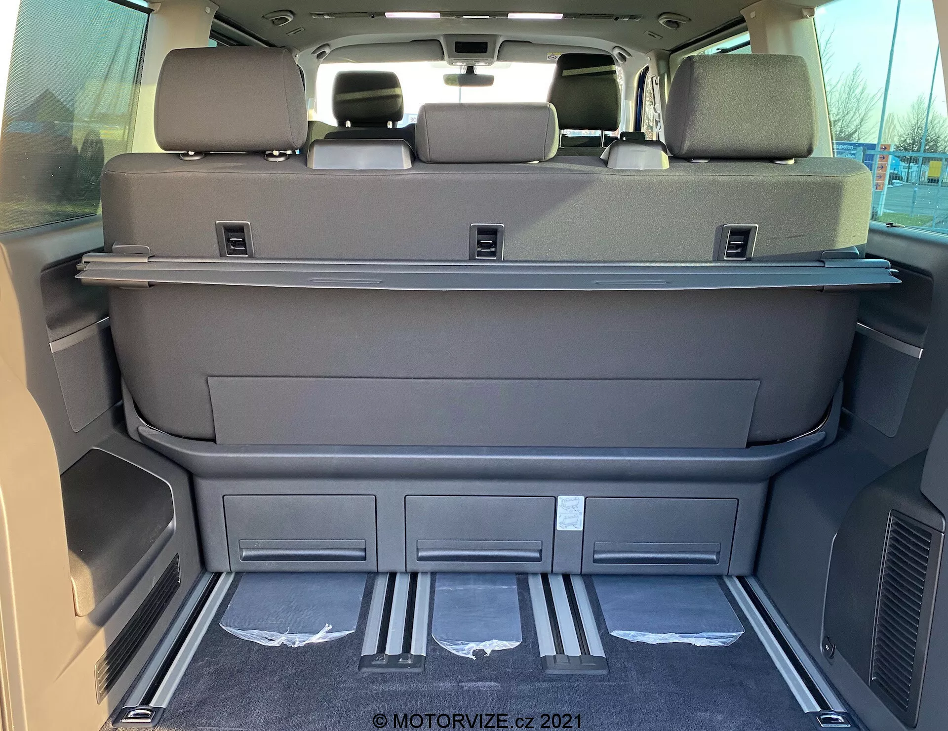 Vista interior do espaço de carga de um VW Nutzfahrzeuge Transporter T6.1 Multivan 2019 a partir da parte traseira, mostrando uma área espaçosa com os bancos da terceira fila rebatidos, cor cinza escuro ou preto, prateleira de cobertura de carga, encostos de cabeça nos bancos traseiros, guardas de entrada metálicas, compartimentos moldados, ventiladores, carpete limpa e possíveis controles eletrônicos dos bancos ou da tampa traseira.
