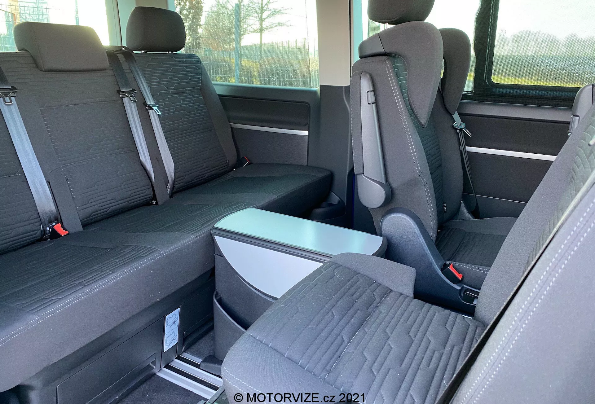 Vista interior do VW-Nutzfahrzeuge Transporter 2019 (T6.1 Multivan) focada nos bancos da segunda e terceira filas. Fotografado de lado ao nível dos olhos, a imagem mostra bancos individuais com apoios de cabeça proeminentes na segunda fila e um banco com capacidade para três passageiros na terceira fila. Os bancos são de tecido bicolor e o piso possui trilhos de metal para assentos reconfiguráveis. Um apoio de braço dobrável e fivelas de cinto de segurança são visíveis. A janela e o armazenamento lateral também são parcialmente visíveis.