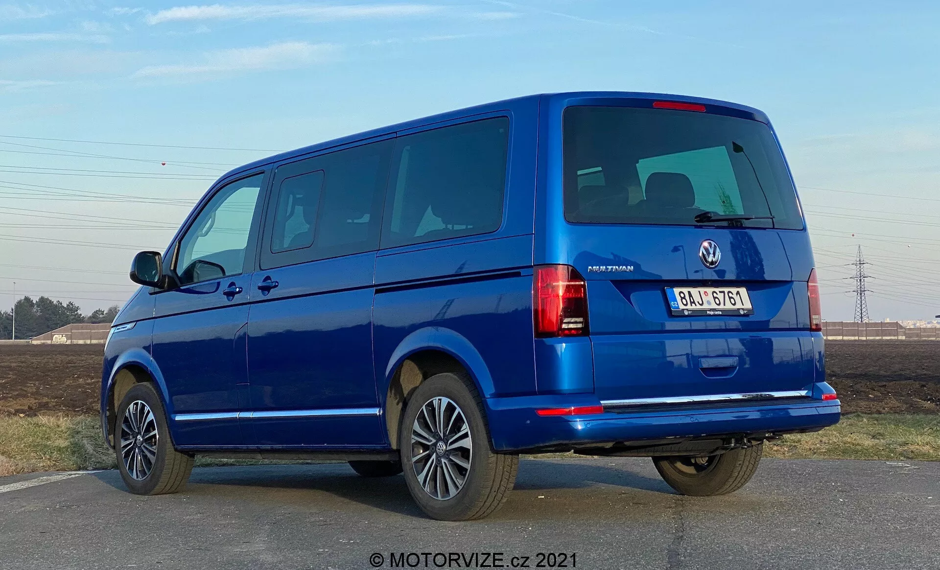 Uma van azul VW-Nutzfahrzeuge Transporter 2019 (T6-1 Multivan) vista de um ângulo traseiro de três quartos. A imagem captura os conjuntos de luzes traseiras, janelas traseiras e porta-malas, bem como o perfil lateral com portas e janelas de passageiros. A van ocupa uma grande parte da moldura ao nível dos olhos, revelando portas traseiras divididas e várias janelas laterais, indicando um modelo de passageiros. Rodas de liga leve, para-choques da cor da carroçaria e espelhos laterais sugerem um alto nível de acabamento. A iluminação sugere durante o dia com céu claro, e há sombras nítidas debaixo do veículo.