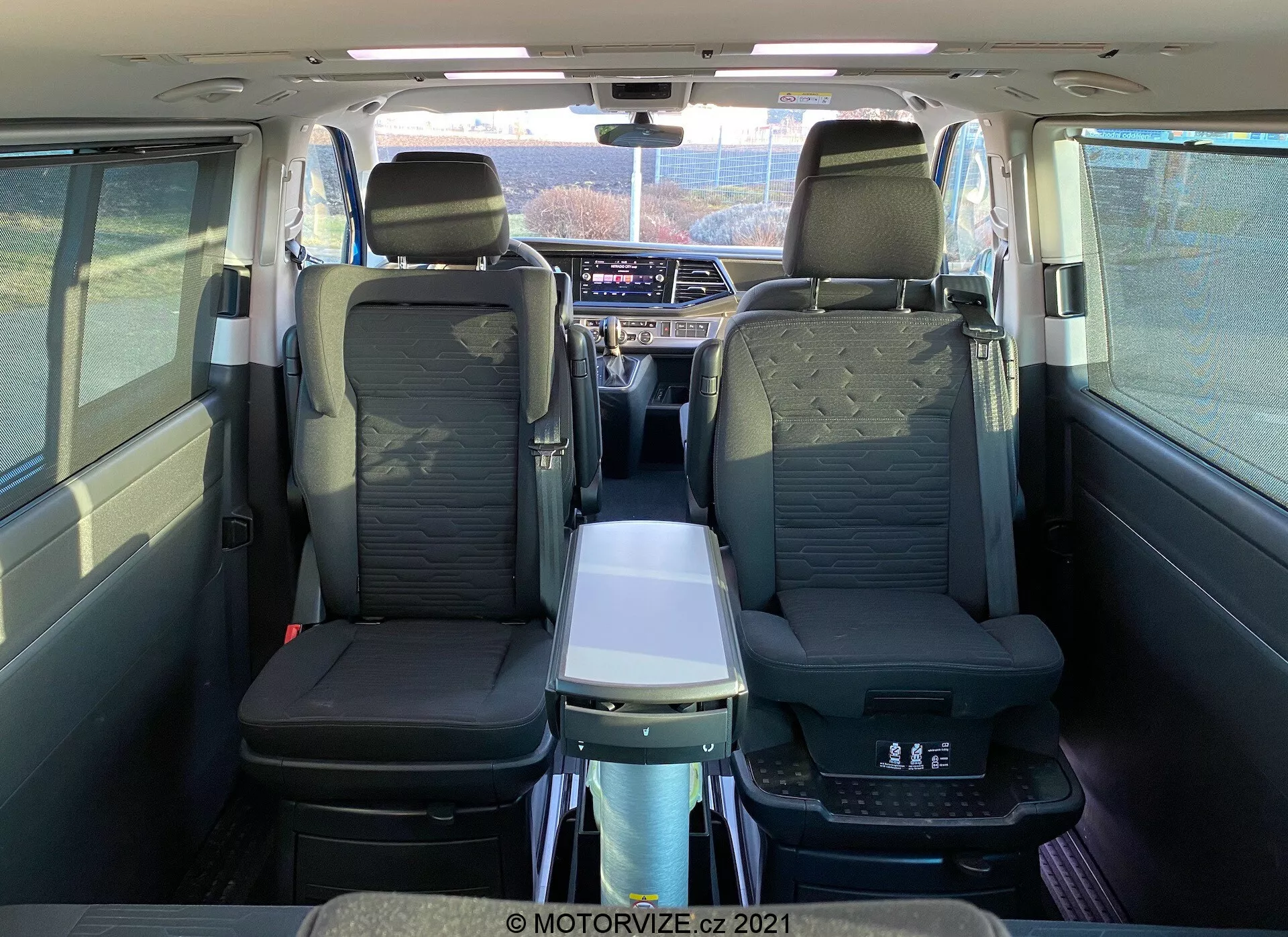 Interior do VW Nutzfahrzeuge Transporter T6-1 Multivan 2019 visto de trás, mostrando assentos de couro, apoios de braço na segunda fila, mesa dobrável, teto panorâmico, saídas de ar individuais e avançado sistema de infoentretenimento; destaca o conforto dos passageiros e a cabine espaçosa.