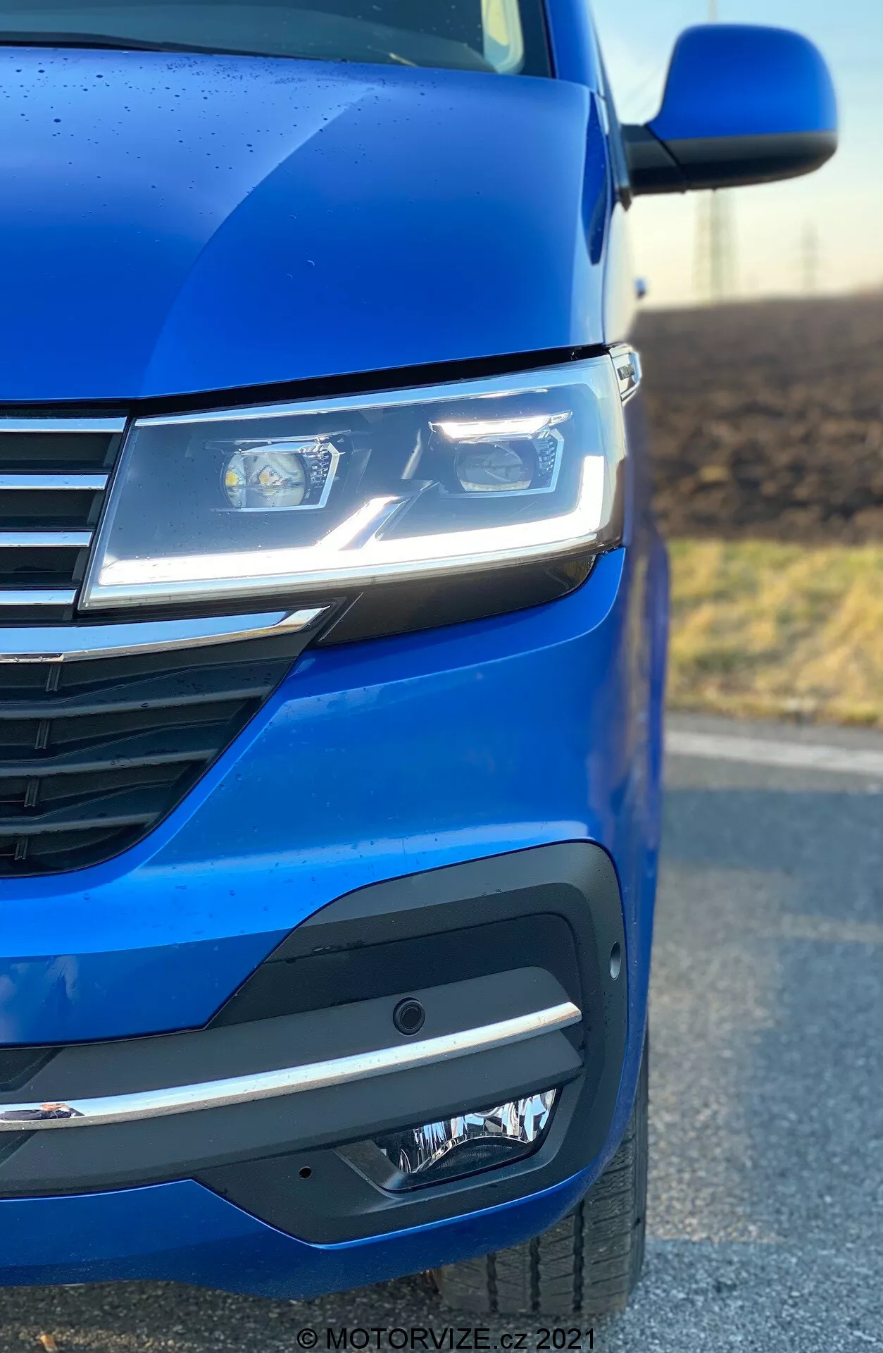 Close-up do lado frontal esquerdo de um VW Nutzfahrzeuge Transporter 2019 (T6.1 Multivan) destacando a pintura azul metálica, faróis LED avançados, design moderno da grade com linhas horizontais e um sofisticado para-choque dianteiro com luzes de neblina ou unidades de sensores. O ângulo da foto enfatiza a aerodinâmica e os detalhes de design do carro.