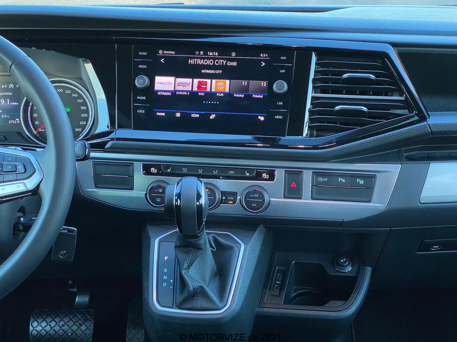 Visão do interior do VW Nutzfahrzeuge Transporter 2019 T6.1 Multivan a partir da perspectiva do assento do passageiro, mostrando o painel moderno e o console central. Volante multifuncional revestido em couro com controles de mídia, grande sistema de infoentretenimento com tela sensível ao toque mostrando opções de mídia e conectividade, saídas de ar elegantes, botões táteis para funções rápidas, painel de controle de clima de dupla zona, área ergonômica da alavanca de câmbio automático e botão de partida/parada sem chave. O design combina plásticos escuros, acabamentos que lembram metal escovado e materiais semelhantes ao couro, destacando um ambiente contemporâneo e premium.