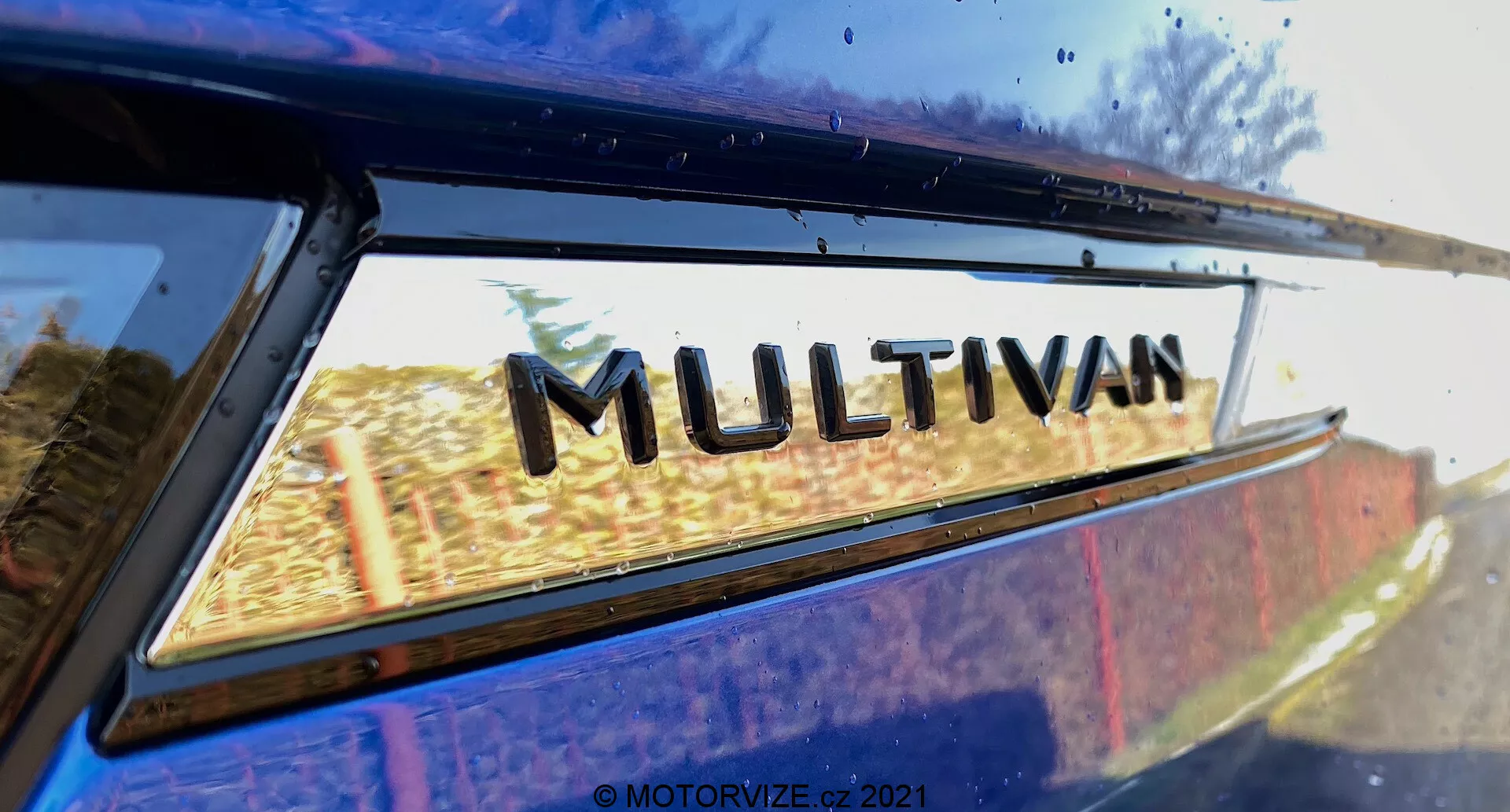Visão aproximada de uma parte do VW Nutzfahrzeuge Transporter 2019 (T6-1 Multivan) com foco no emblema 'MULTIVAN'. A imagem é tirada de um ângulo traseiro baixo, olhando ligeiramente para cima onde a janela traseira se encontra com a borda do teto. Leves reflexos no vidro da janela sugerem um cenário ao ar livre. Esta imagem recortada de perto enfatiza o emblema de designação do modelo do fabricante.