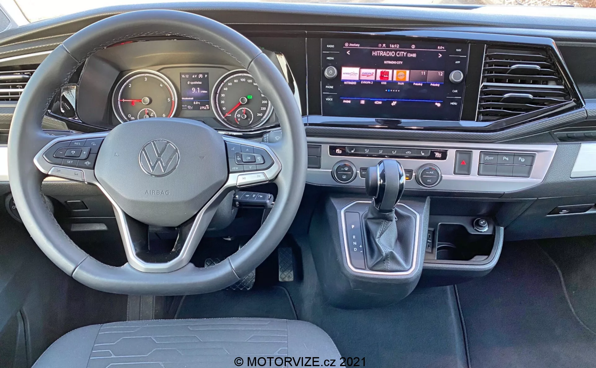 Vista interior do VW Nutzfahrzeuge Transporter 2019 T6.1 Multivan da perspectiva do assento do motorista. Características: volante com a marca Volkswagen e botões multifuncionais, medidores analógicos, display digital, console central com sistema de infotainment touchscreen ativo, controle de clima de dupla zona, alavanca de câmbio com botão de estacionamento eletrônico e acentos modernos em metal escovado.