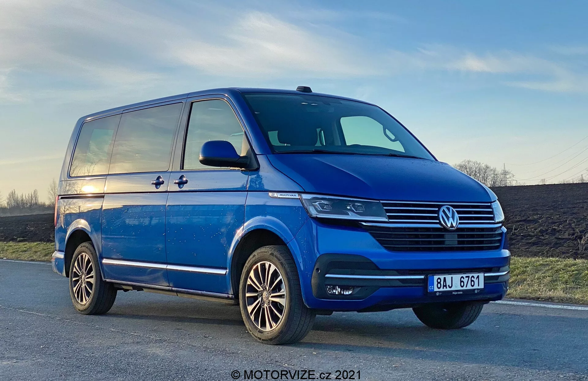 Vista de baixo ângulo de uma Volkswagen T6-1 Multivan azul 2019, mostrando o quadrante dianteiro esquerdo de uma perspetiva três-quartos. A carrinha está estacionada numa estrada ligeiramente inclinada, com detalhes cromados visíveis na grelha, um emblema VW central e unidades de faróis modernos que podem utilizar tecnologia LED. Os faróis de nevoeiro estão integrados no para-choques, que também possui entradas de ar. O perfil lateral revela uma porta deslizante, uma linha de caráter que se estende do pára-lama dianteiro para a traseira e espelhos retrovisores substanciais com indicadores de direção. As rodas de liga leve com um design de vários raios e o piso dos pneus bem conservados também são visíveis. O céu no fundo mostra um toque de pôr do sol.