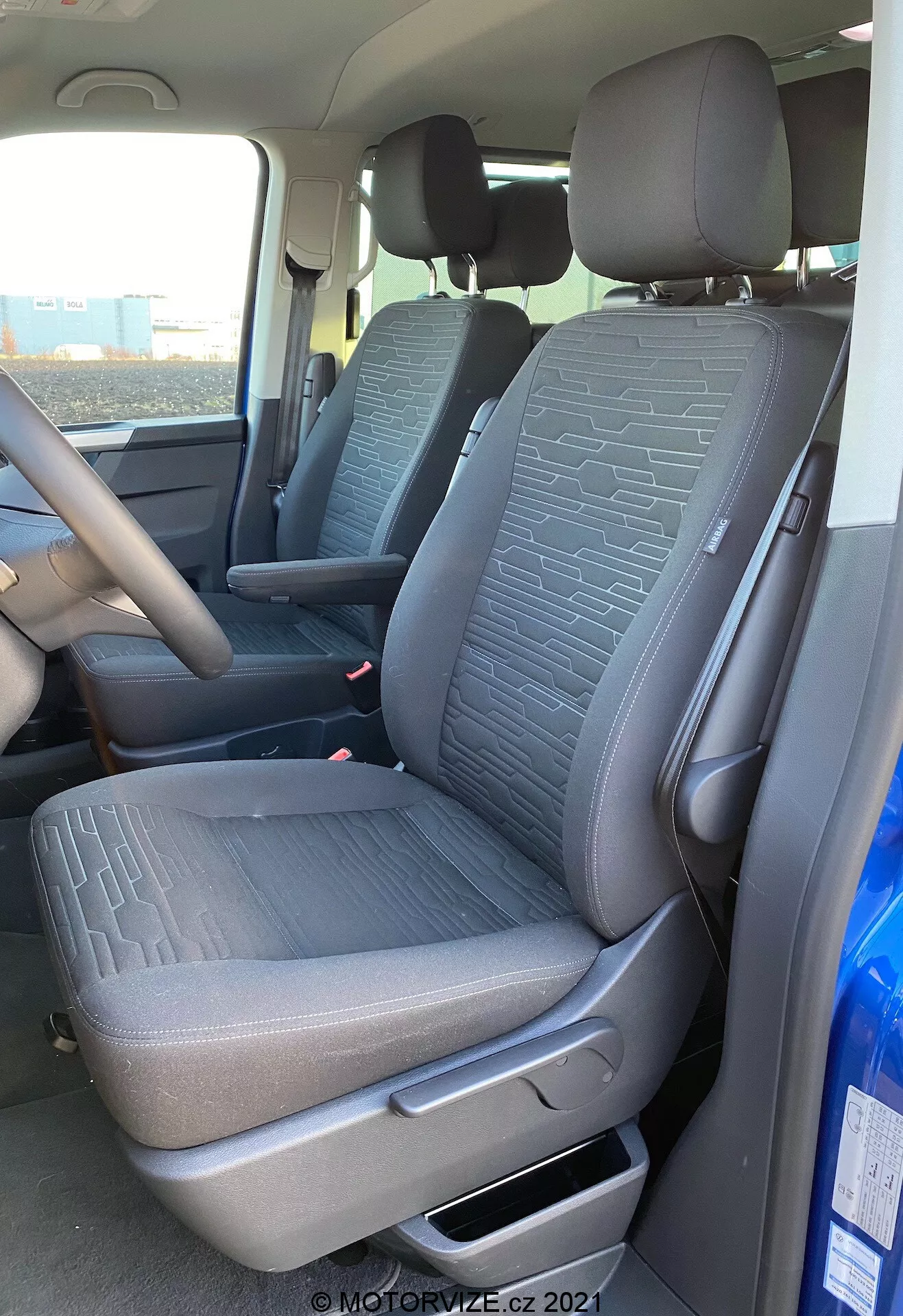 Interior do VW Nutzfahrzeuge Transporter 2019 T6.1 Multivan, mostrando o assento dianteiro do passageiro e parte do assento do motorista a partir de uma perspectiva externa do lado do passageiro. A foto, tirada de um ângulo ligeiramente baixo, destaca o tecido texturizado com padrões geométricos nos assentos, a alavanca de ajuste manual, os cintos de segurança e parte do console central e do volante. A luz natural ilumina o interior do veículo.
