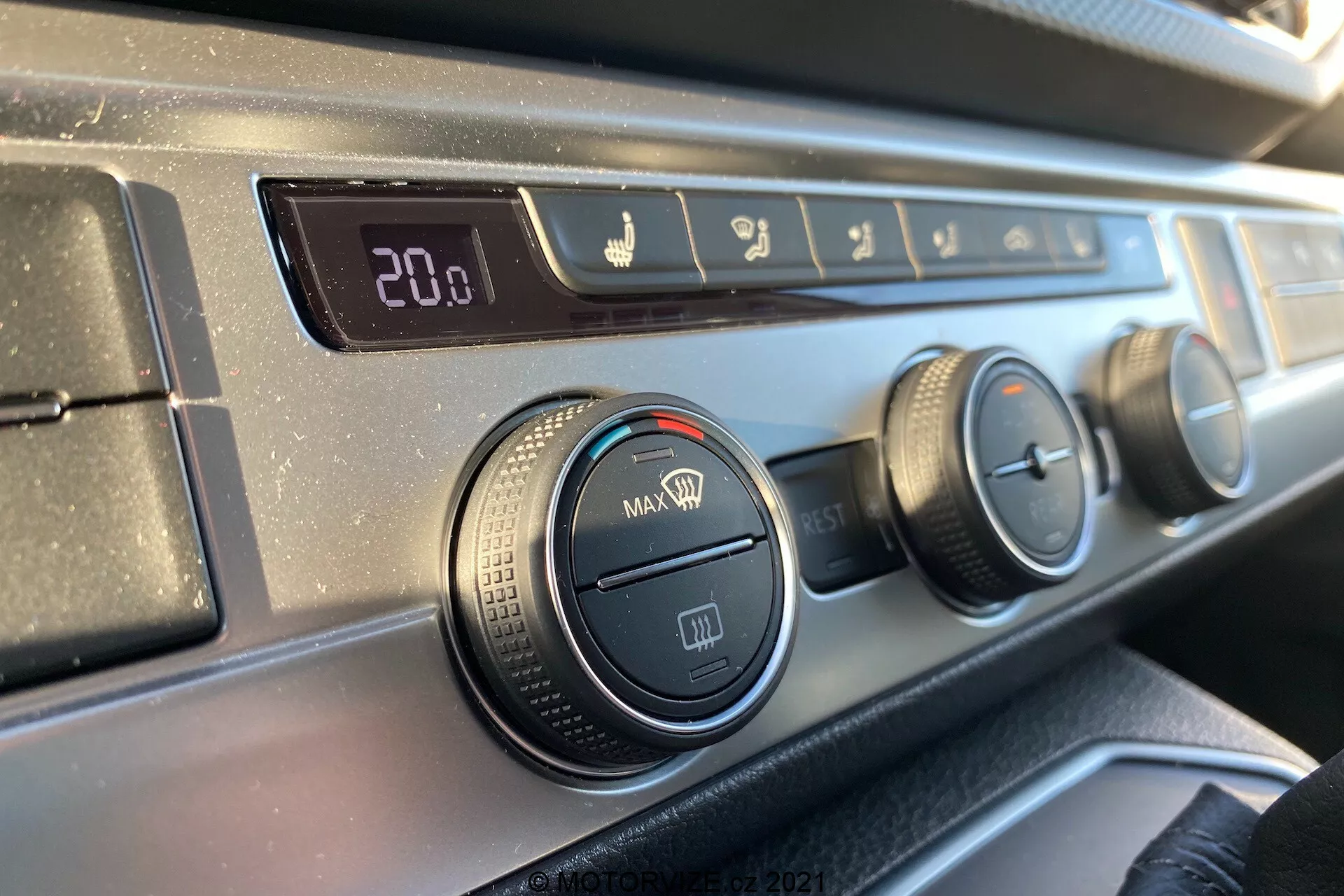 Vista aproximada do painel de controle de climatização no painel do VW Transporter T6.1 Multivan 2019 a partir da perspectiva de um ocupante do assento dianteiro, com um display digital de temperatura ajustado para 20,0 graus Celsius, controles de direção do fluxo de ar, três botões rotativos para ajustes de ar condicionado e ventilador e um botão de recirculação, todos com uma borda de plástico texturizado preto e um acabamento liso e escuro no painel.