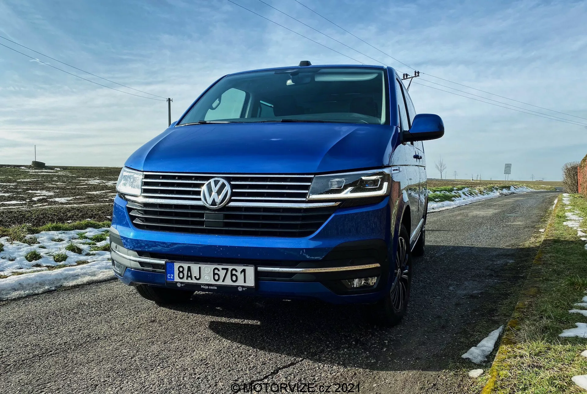Uma vista de três quartos frontal baixa do Volkswagen T6.1 Multivan 2019, apresentando a grade frontal com linhas horizontais, o distintivo emblema VW centrado na grade, faróis possivelmente com luzes diurnas de LED e luzes bi-xenon ou LED, e para-choques dianteiro com luzes de nevoeiro e entradas de ar. A imagem mostra parte do lado esquerdo, incluindo a porta do motorista e o para-lama, com o capô e o para-brisa visíveis. O carro é azul profundo e está estacionado em uma estrada rural com neve no chão e um céu azul claro acima.