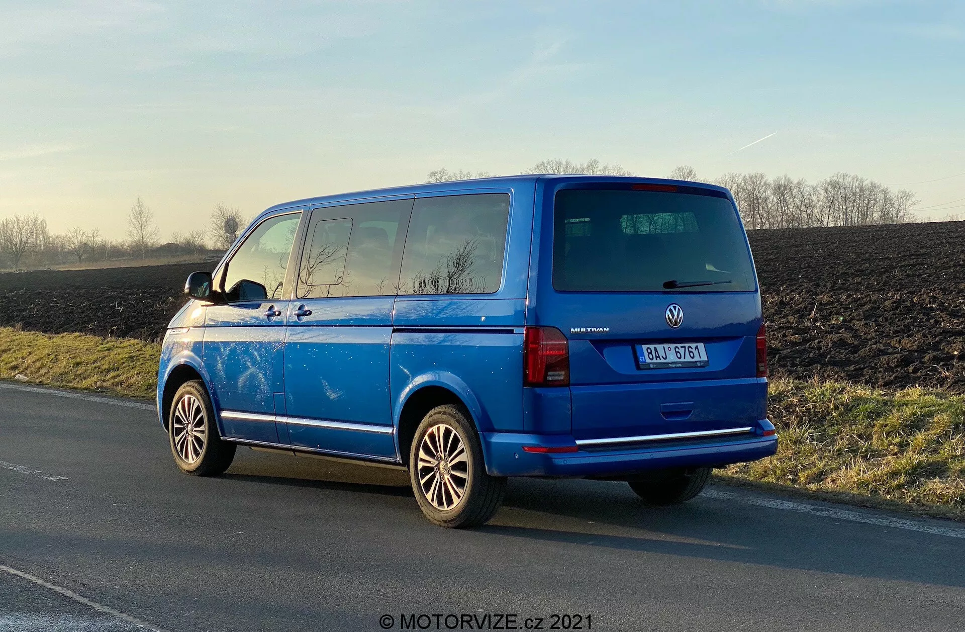 Vibrante Volkswagen Transporter 2019 (T6-1 Multivan) azul estacionado no acostamento da estrada, vista traseira de três quartos mostrando luzes traseiras, janela traseira, porta traseira, emblema do fabricante, detalhes do modelo do veículo; rodas de liga visíveis, espelhos laterais externos, linhas do desembaçador do vidro traseiro, vidros escurecidos da segunda fila, antena no teto; estacionado em luz natural do dia com reflexos destacando a superfície polida, fundo rural.
