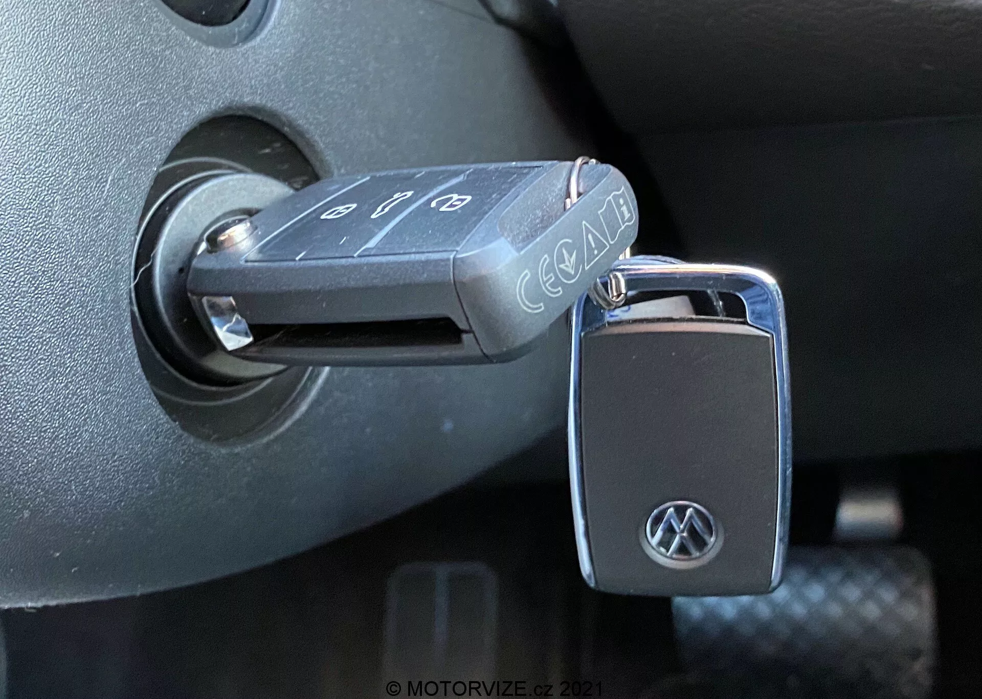 Close-up de uma chave dobrável da Volkswagen inserida no interruptor de ignição de um VW Nutzfahrzeuge T6-1 Multivan 2019. A chave, com botões para travar, destravar e alarme de pânico, exibe um logotipo da VW no chaveiro. O interruptor de ignição está localizado na coluna de direção de plástico escuro, indicando que a ignição está na posição 'desligado'. A foto, tirada de um ângulo lateral com luz natural, destaca as texturas e acabamentos detalhados do interior do veículo.