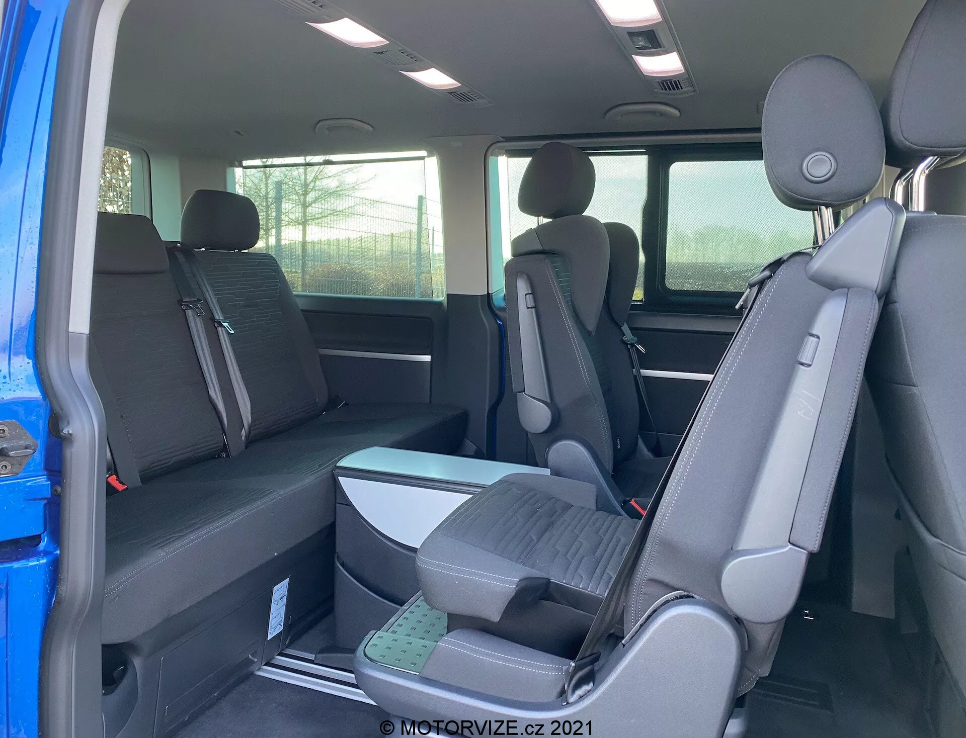 Interior do VW Transporter T6.1 Multivan 2019 mostrando a segunda e terceira fila de assentos a partir da perspectiva do lado do passageiro traseiro, com configuração de assentos 2-3-2 e estofamento em tecido escuro. Vista parcial do exterior através da porta aberta, apoios de cabeça em todos os assentos e assentos elevados na terceira fila para melhor visibilidade.