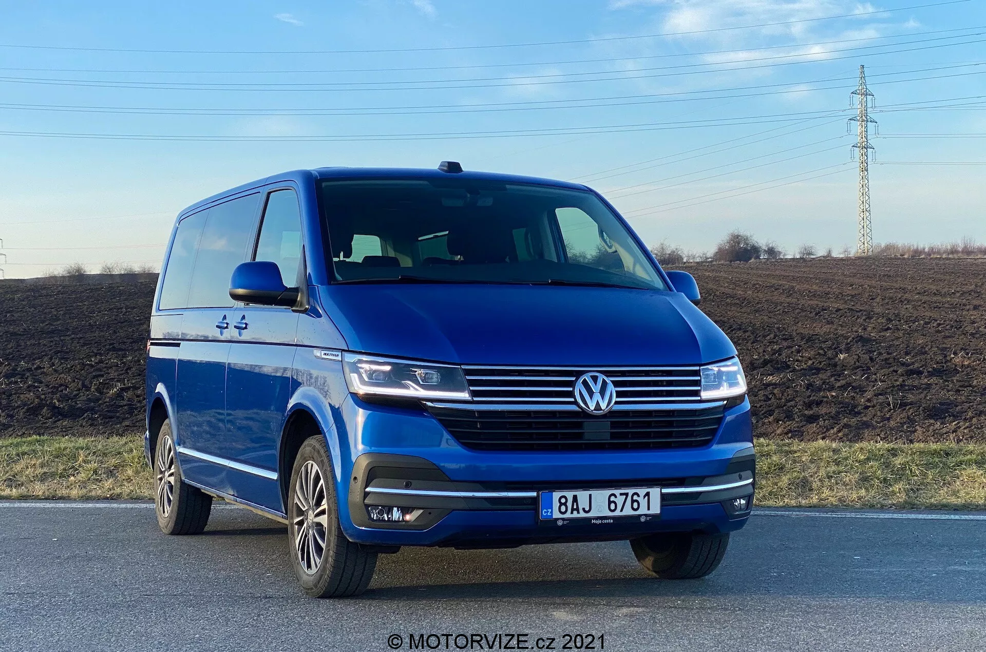 A imagem apresenta uma van azul Volkswagen VW-Nutzfahrzeuge Transporter 2019 (T6-1 Multivan) capturada de um ângulo frontal baixo, destacando o quarto frontal esquerdo com uma visão clara da sua fachada frontal e perfil do lado do motorista. O veículo está em uma estrada, destacando elementos como o distintivo proeminente da Volkswagen, faróis elegantes, linhas esculpidas no capô, faróis de neblina integrados no para-choque e rodas de liga leve de múltiplos raios. A fotografia, tirada à luz do dia, enfatiza o design exterior da van e tecnologias de assistência ao motorista, como os sensores de estacionamento.