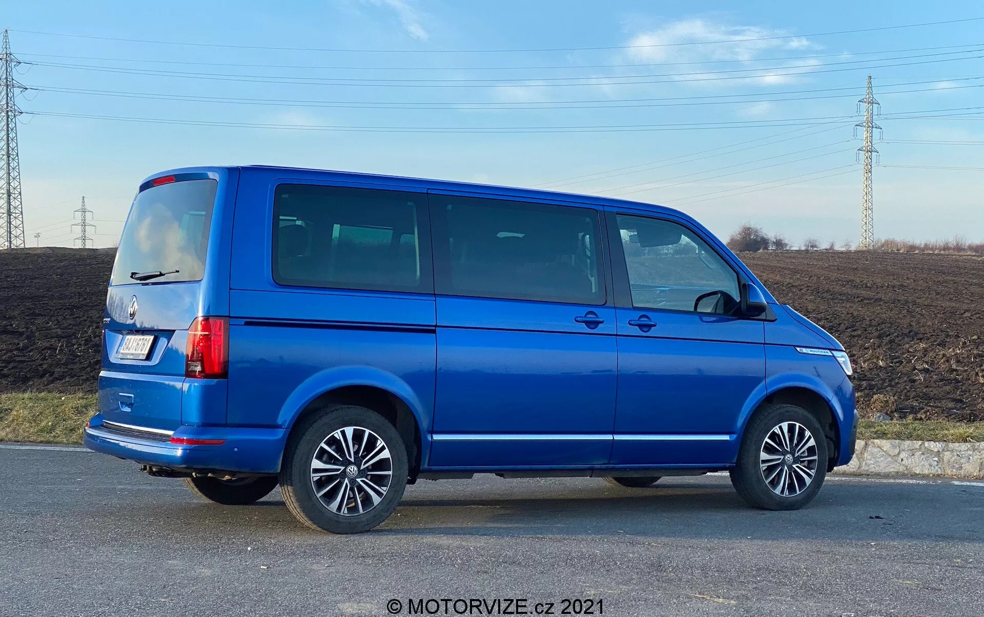 Vista completa do perfil lateral de um VW Nutzfahrzeuge Transporter azul de 2019 (T6.1 Multivan) com rodas de liga, linhas de porta visíveis, janelas laterais escurecidas, silhueta quadrada, luzes traseiras verticais, antena no teto e pintura metálica azul brilhante. A imagem foi tirada em um ângulo, proporcionando perspectiva tanto do lado quanto da parte traseira do veículo, situado em uma área rural com um campo e linhas de energia ao fundo, em luz diurna.