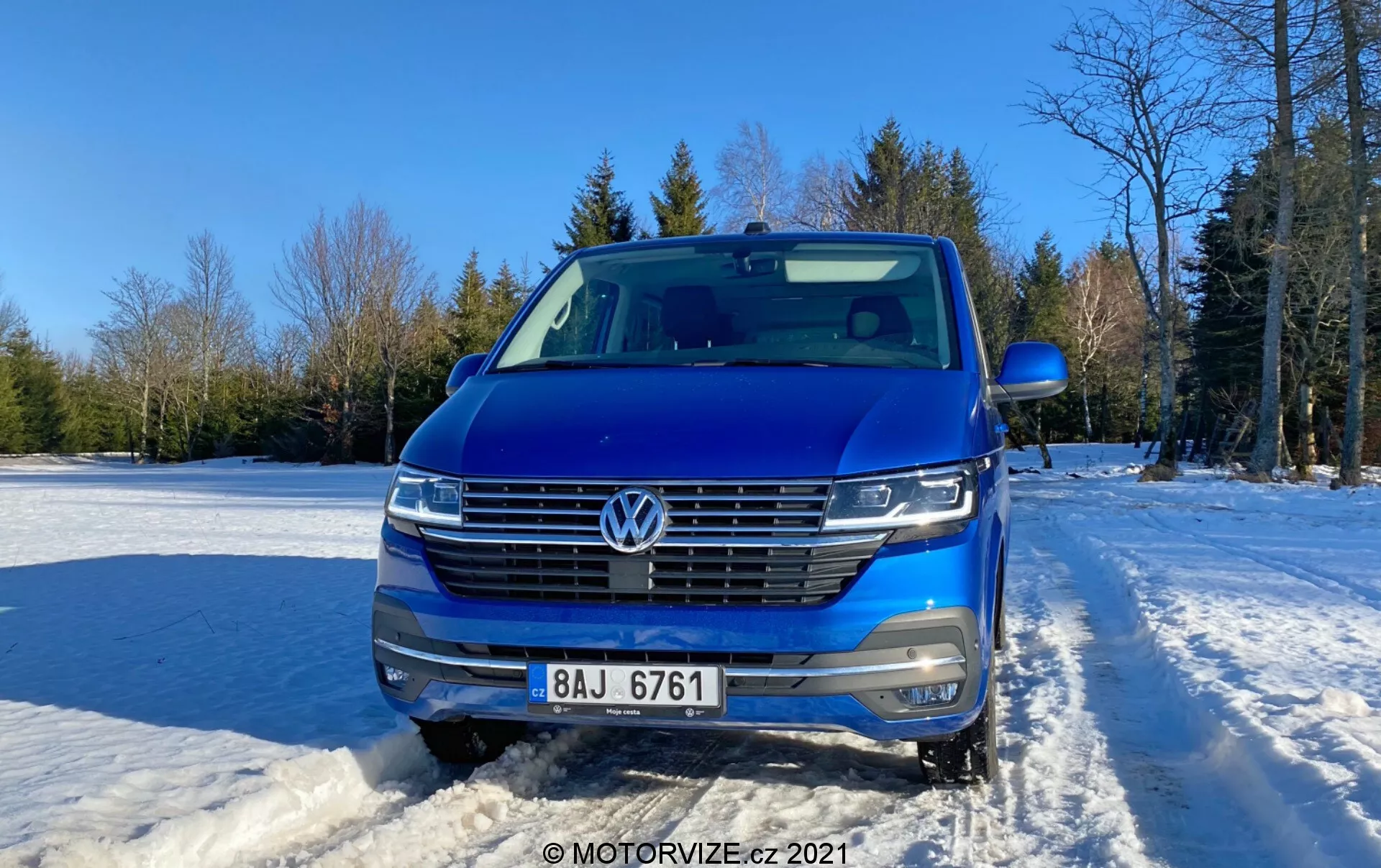 Foto frontal de um Volkswagen Transporter 2019 (T6-1 Multivan) azul, mostrando sua grade, faróis e área do capô. Posicionado ao ar livre em solo coberto de neve com céu claro e árvores ao fundo. As características do carro incluem emblema VW, grade horizontal com acabamento cromado, faróis LED, câmera frontal, sensores de estacionamento, trilhos de teto prateados e espelho lateral parcialmente visível. Design contemporâneo com frente aerodinâmica, fotografado durante o dia com luz suave do sol a partir do lado direito.