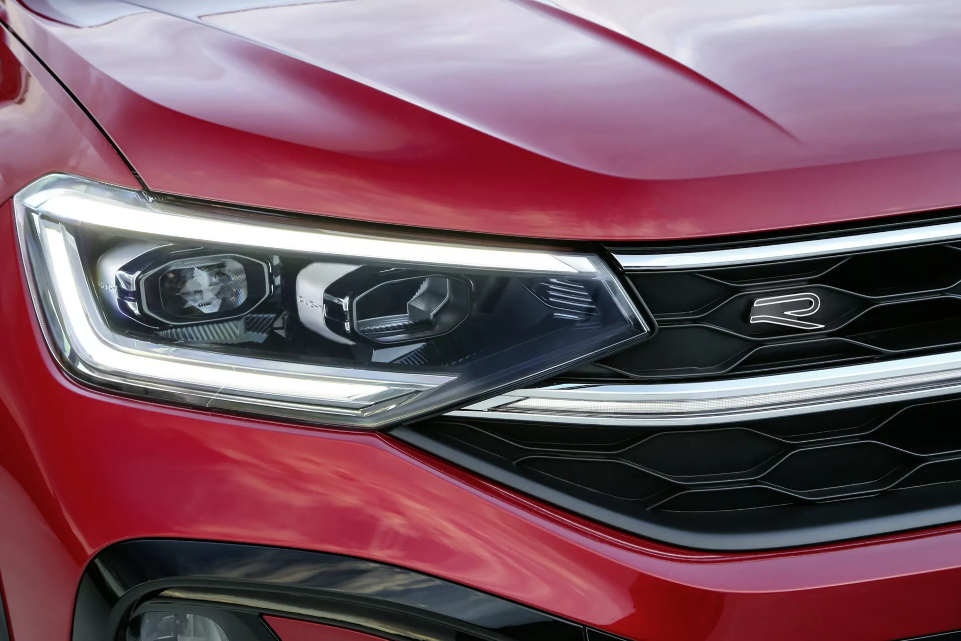 Close-up de um VW Taigo 2021 vermelho, destacando o farol esquerdo, para-choque dianteiro e parte da grade com acabamentos em cromo, exibindo iluminação LED, design moderno e aparência esportiva e de luxo