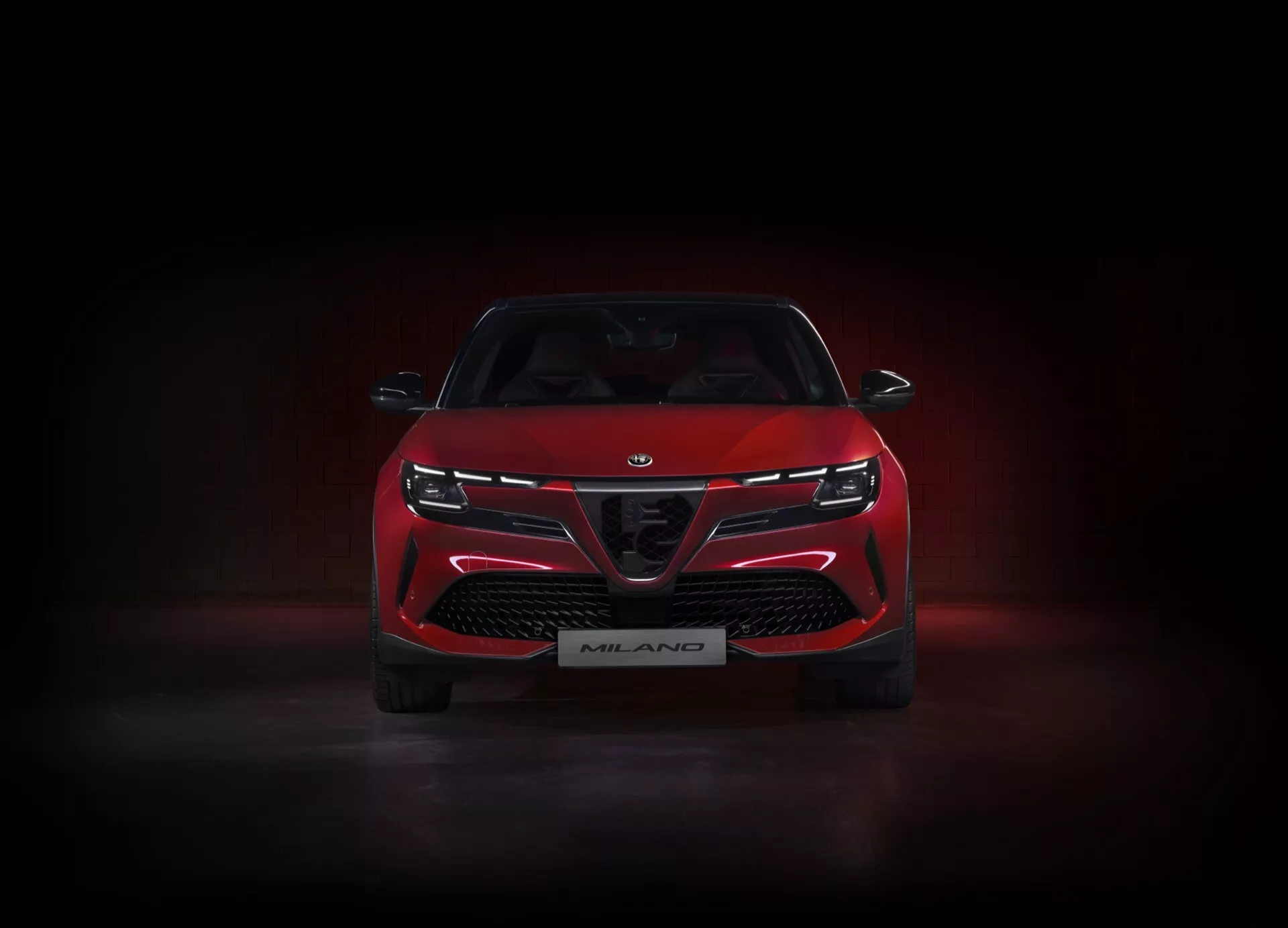 Vista frontal do Alfa-Romeo Junior 2024 (966) em vermelho, iluminado em estúdio contra um fundo escuro com um brilho sutil por baixo, destacando sua grade distintiva com grandes entradas de ar, faróis LED estreitos e elegantes conectados por uma faixa de luz, e emblema da edição Milano, enfatizando suas características de design esportivo e agressivo.