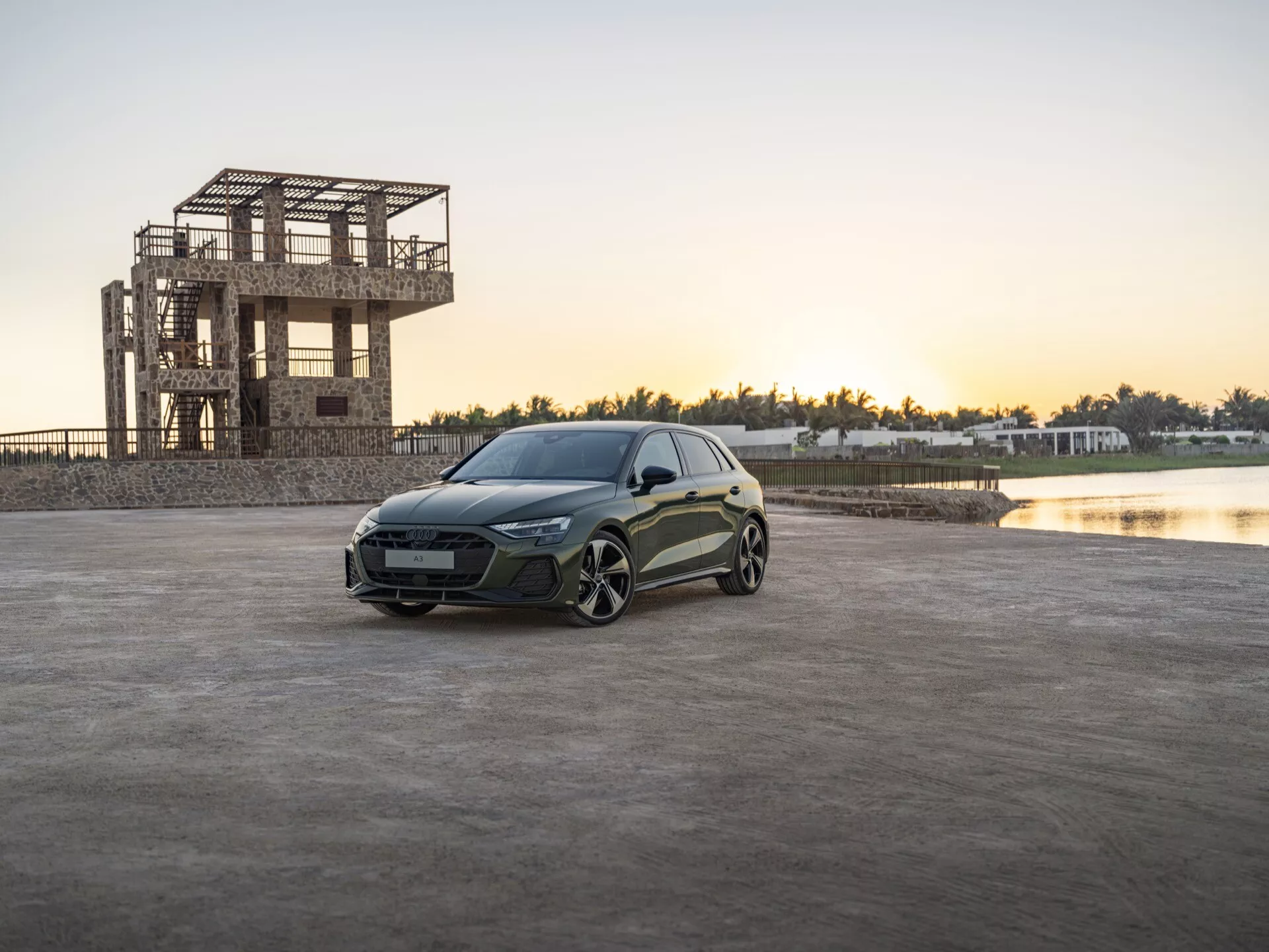 Audi A3 (8Y-Facelift) 2024 posicionado em uma área pavimentada aberta ao amanhecer ou entardecer com um prédio em construção e um corpo d'água ao fundo, exibindo os faróis LED elegantes, a carroceria esculpida e as rodas de liga leve orientadas para desempenho do veículo.