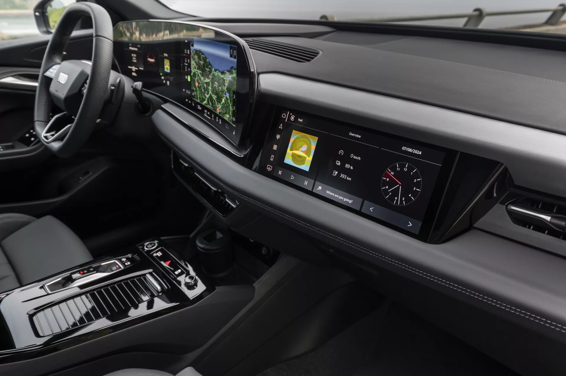 Vista interior do Audi Q6 e-tron 2024 focando no painel de bordo e na consola central a partir de um ângulo ligeiramente elevado. Do lado esquerdo, o volante, indicando uma configuração para condução à esquerda, enquanto a consola central apresenta uma alavanca de mudanças, vários controles e um grande ecrã tátil. O painel de instrumentos digital exibe métricas de condução. O painel de bordo estiloso estende-se horizontalmente com aberturas de ventilação e outro elemento de design à direita. A tecnologia avançada é evidente com o ecrã tátil central exibindo informações de navegação e multimídia, um head-up display visível acima do volante e botões sensíveis ao toque para o controle climático. O design enfatiza o minimalismo e materiais de alta qualidade, com costuras de couro e detalhes metálicos que adicionam à sensação de luxo.